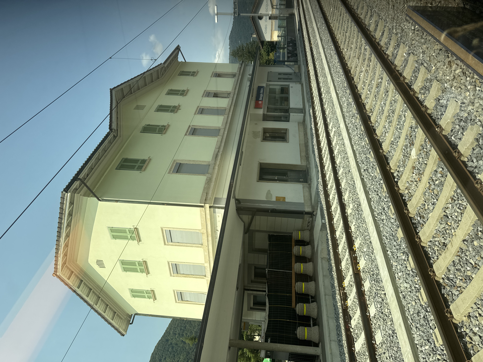 This is a photograph taken from a train, showing the train station in Maroggia, a village within the municipality of Val Mara, Switzerland. The image captures a light green, three-story building, which appears to be the main station building. It has a sloping roof with multiple dormer windows on the upper levels and green shutters on the windows. Beneath the main building is a covered platform with several stone bollards topped with yellow rings lined up along the edge. Railway tracks run in the foreground, leading towards the station. To the right of the main building, a smaller, white building with "casa dell'acqua" written on it is visible. The scene is set against a backdrop of green, tree-covered hills and a clear blue sky with some clouds. The photograph is taken in daylight.