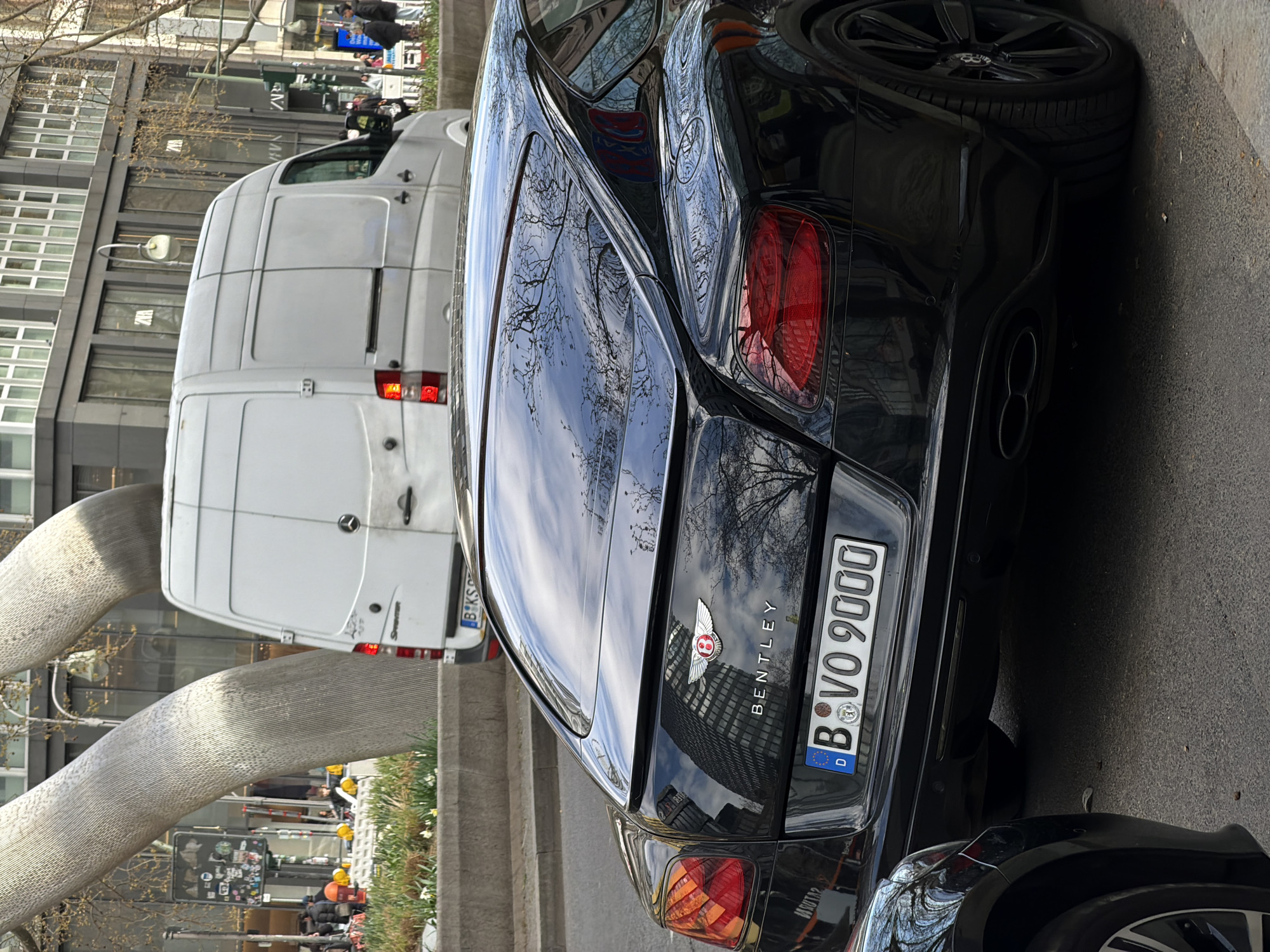 A black Bentley, license plate "B VO 9000", is parked on a street in Berlin, Germany. A white Mercedes Sprinter van is parked next to it. The license plate of the van is partially visible, "B-KS". There's a large metallic sculpture of interconnected tubes in the background and building reflected on the trunk of the car. Pedestrians and traffic are visible in the background.