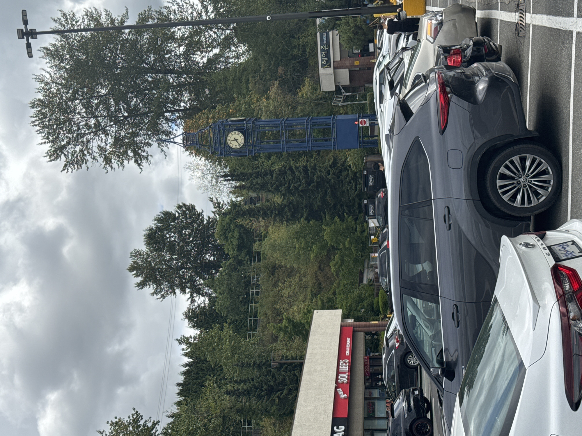 The image captures a scene in Coquitlam, Canada, likely a commercial area with a parking lot. In the foreground, several cars are parked, including a white Toyota Camry and a dark gray SUV. The SUV has a distinctive rear light design. In the middle ground, a blue clock tower, reminiscent of Big Ben, stands tall, featuring a clock face with Roman numerals. Adjacent to the clock tower is a building with signage that includes Korean characters and the name "HairZone." Further to the left, another building bears the sign "SOL LEE'S KOREAN RESTAURANT." The background is dominated by a hillside covered in lush green trees, indicating a natural landscape bordering the urban development. The sky is overcast with gray clouds, suggesting a cloudy day. A tall lamppost with multiple lights and a security camera is visible on the right side of the image. Two individuals are partially visible in the image, one standing near the dark gray SUV and another near the building with the "HairZone" sign.