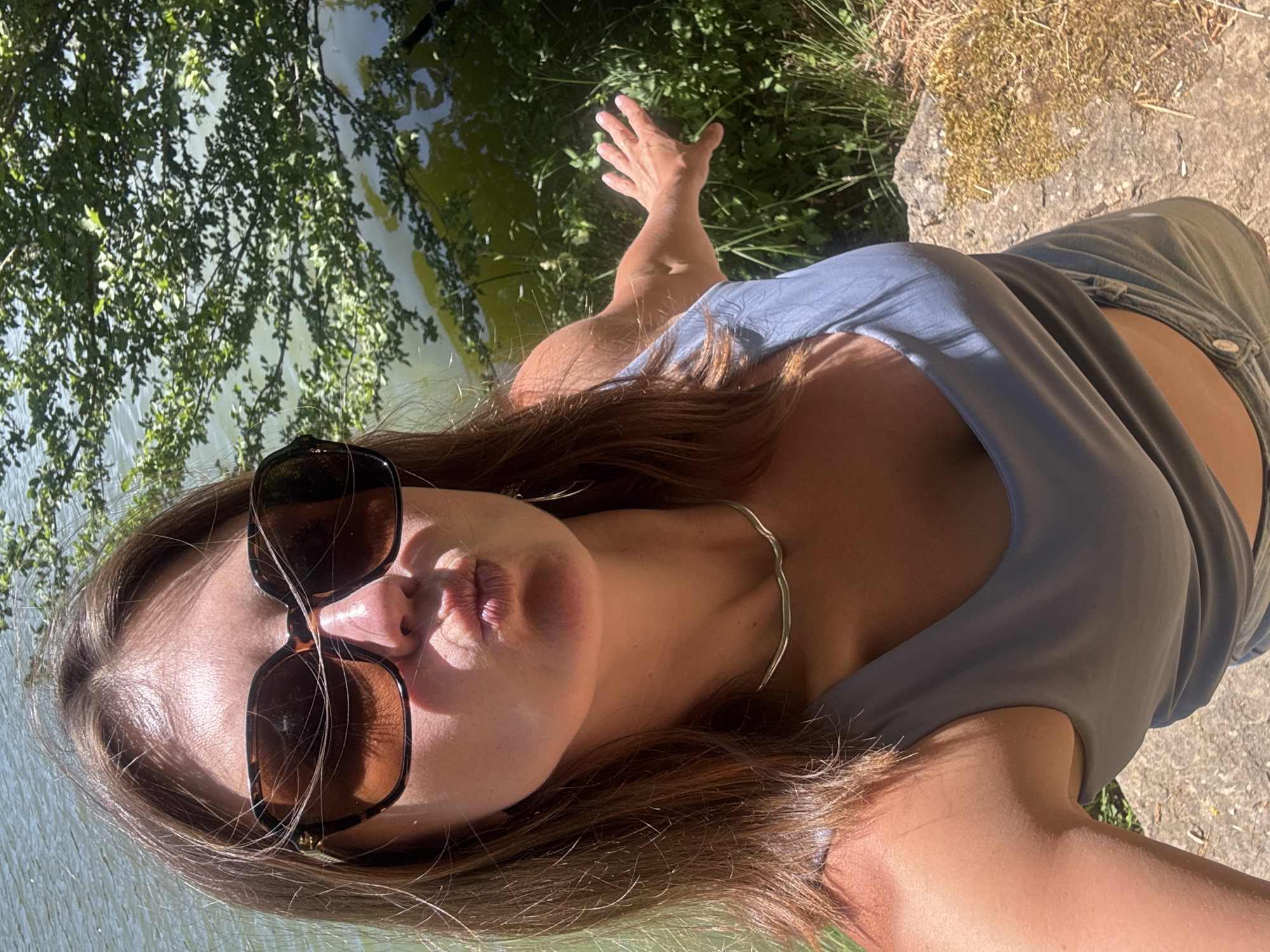 A woman is taking a selfie near a body of water surrounded by trees. She is wearing sunglasses, a blue tank top, and denim shorts. Her right arm is extended to her side, with her hand open. The background shows a body of water with reflections of trees and foliage. The ground in the foreground is rocky with dry grass. The location is stated to be in the city of Ravilloles, France.