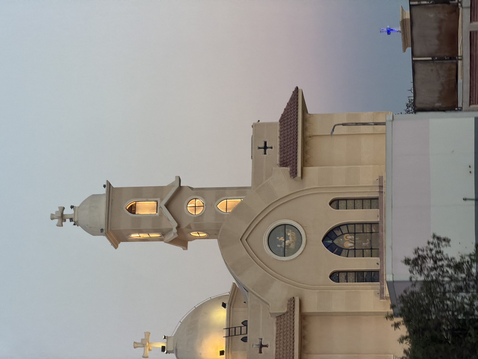A close-up, vertical shot captures the upper portion of a beige, modern church building against a pale, dusk sky. The church features a prominent dome on the left, topped with a cross, and a multi-tiered bell tower on the right, also adorned with a cross. Several arched windows are visible on the tower and the main building, some illuminated from within. A circular stained-glass window above the arched windows depicts a scene with figures. A smaller, black cross is visible on a ledge to the right. In the background, a distant, blue, illuminated cross stands on a separate structure, and a section of a metal fence is present in the foreground. Trees are visible at the bottom left, and part of a white and grey wall is in the lower portion of the image. This church is likely the St. Mark Coptic Orthodox Church in Sharm El Sheikh, Egypt, given its architectural style and the prevalence of Coptic churches in the region.