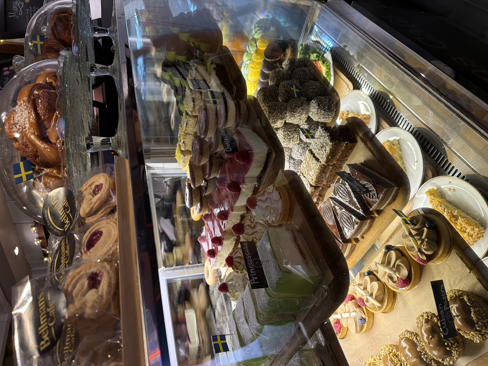 The image shows a close-up, eye-level, interior view of a bakery display case filled with various pastries and cakes. The bakery appears to be "Bellmans Cafe" based on the branding on some of the packaging and the presence of Swedish flags, suggesting a location in Stockholm, Sweden.
The display case is divided into several shelves. The top shelf features large, round pastries under glass domes, one of which is a cinnamon bun topped with pearl sugar and decorated with a Swedish flag. To the left, individually packaged cookies with a jam filling are visible, also bearing the "Bellmans Cafe" logo.
The middle shelf prominently displays several slices of a green marzipan-covered cake, labeled "Princesscake". Next to it are slices of cheesecake with raspberry toppings and small meringue-like desserts. Further to the right, a selection of smaller pastries and what appear to be donuts are arranged.
The bottom shelf contains rows of individual tarts topped with a creamy filling and garnished with berries and a wafer stick. One of these is labeled "Cuban Pastry". To the right of the tarts are slices of chocolate cake.
The overall environment is brightly lit, highlighting the colorful and meticulously arranged desserts. The display case itself is made of glass and metal, with a stainless steel exterior visible at the bottom of the frame. The scene is static, with no people or visible activity.