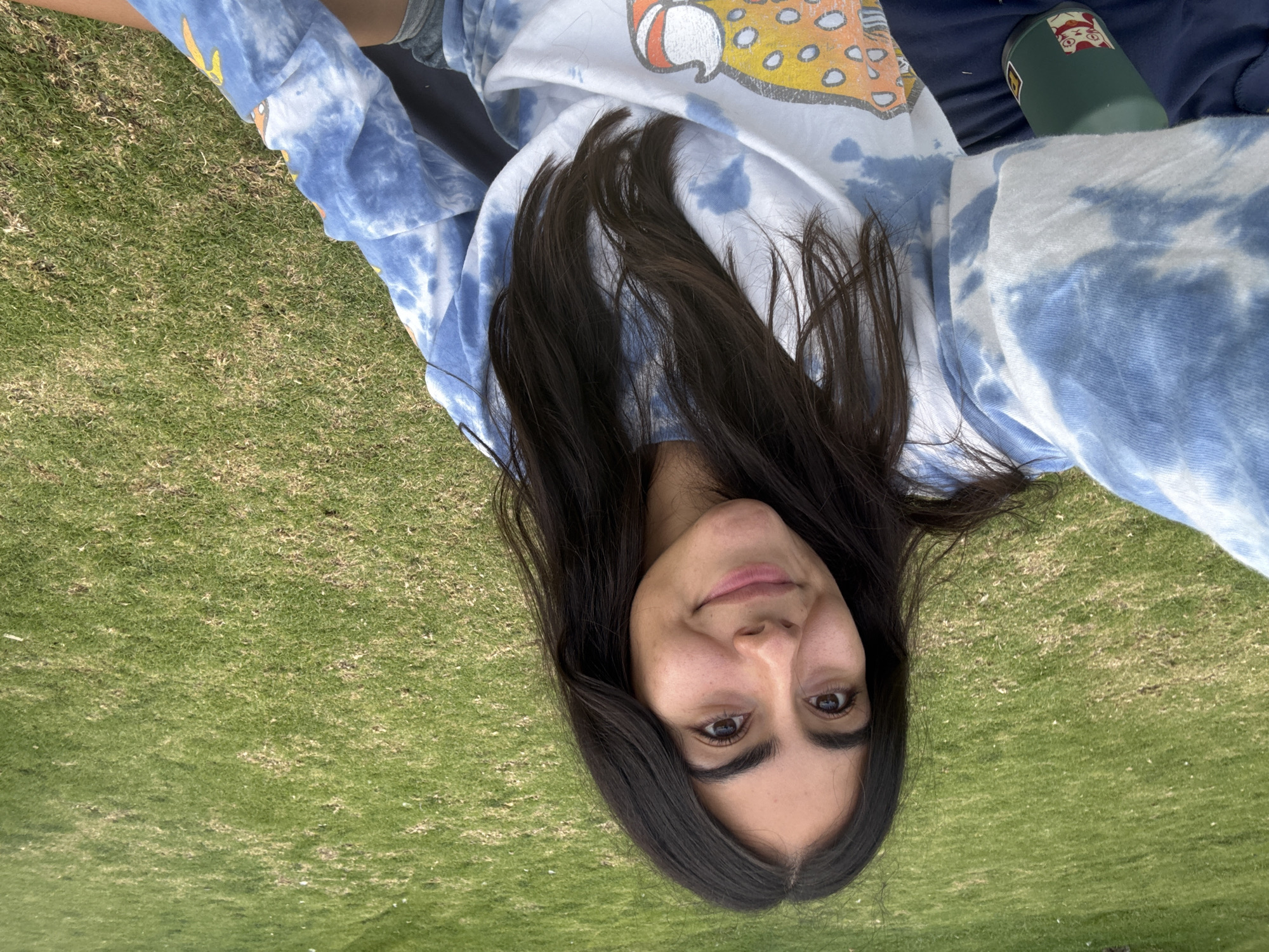 The image shows a young woman with long, dark hair lying on a patch of green grass. She is wearing a tie-dye sweatshirt with a cartoon graphic on the front. To the upper-right, a green water bottle with stickers is partially visible. The location is likely a park or lawn in Puebla, Mexico.