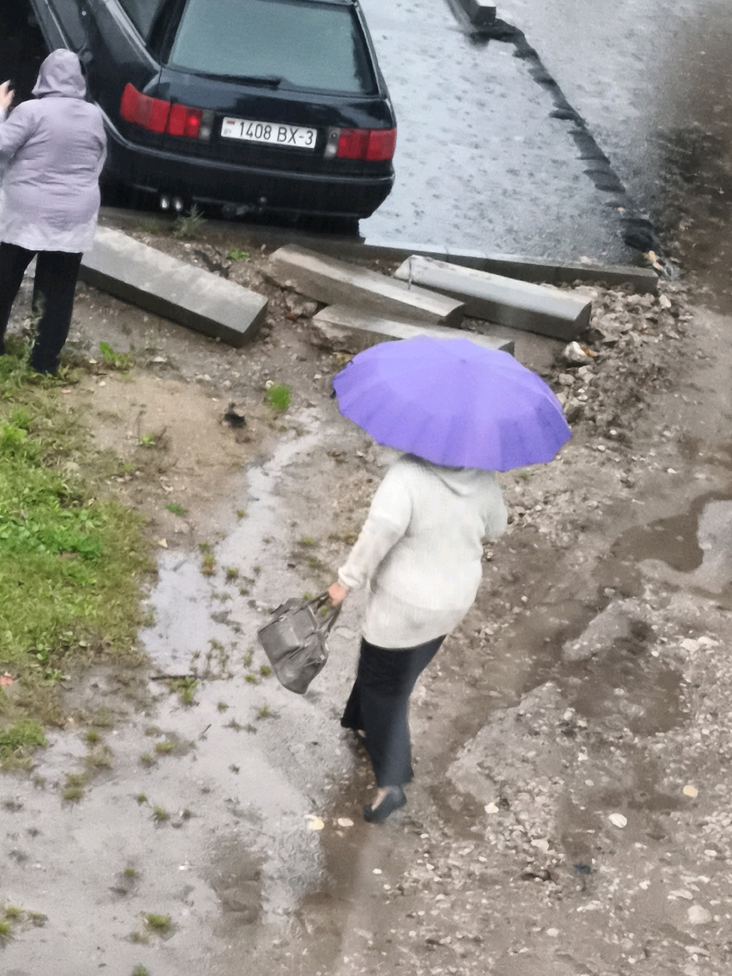 A person is walking away from the camera while holding a purple umbrella. They are wearing a light-colored hooded jacket and dark pants. They are also carrying a dark gray handbag in their left hand. The ground is wet and muddy, with some puddles. There is a black car parked to the left of the frame. The car's license plate is visible and reads "1408 BX-3". The scene appears to be in an outdoor urban or semi-urban environment, potentially during a rainy day. The location is identified as Горскі сельскі Савет, Belarus.