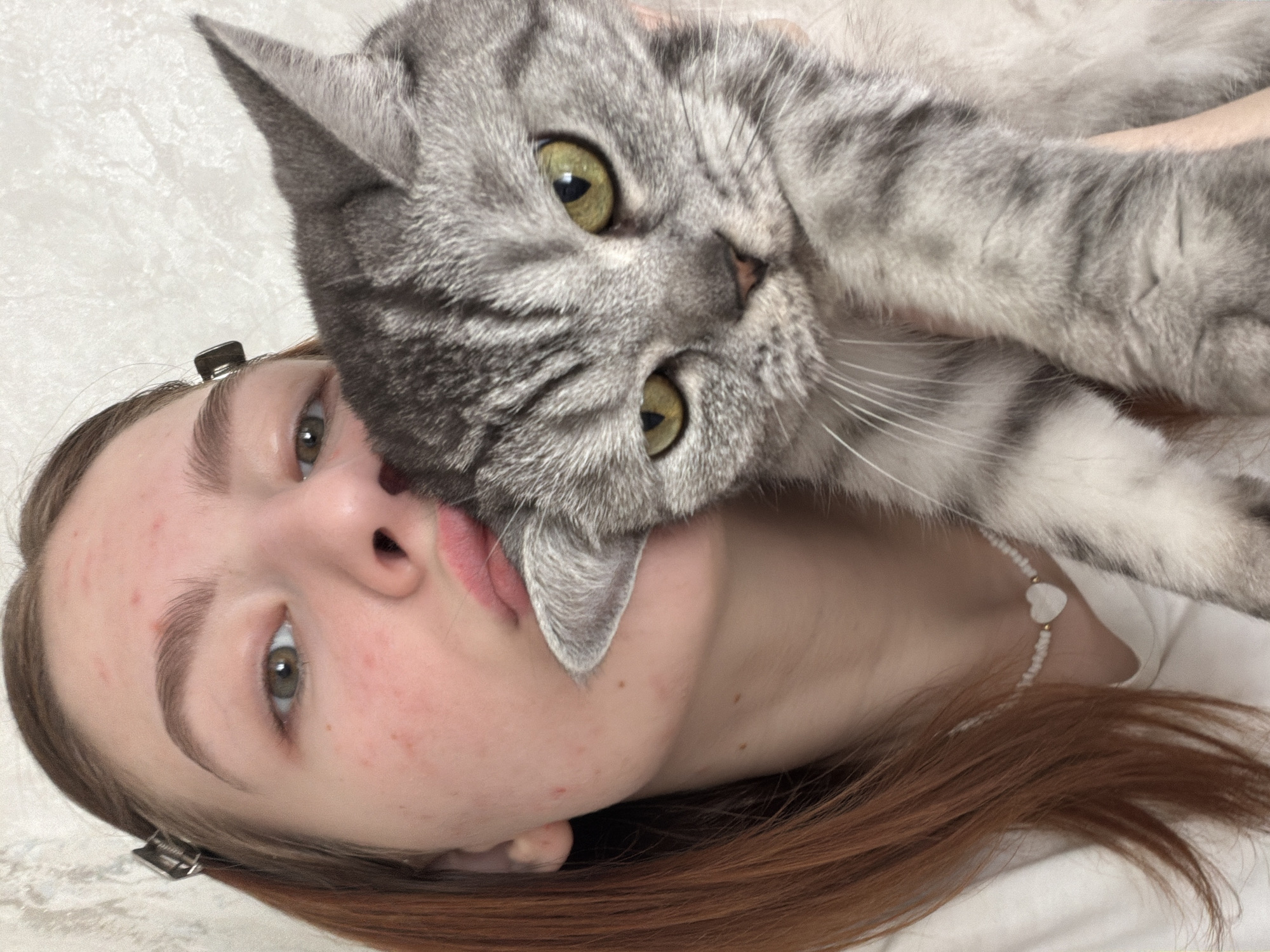 A photograph shows a light-skinned woman and a gray tabby cat. The woman is lying on her back, facing slightly to the left, with her head tilted. The cat is positioned on top of her, with its face close to hers, and the cat's ear touching the woman's lip. The woman has light brown hair, pale skin, and green eyes. Her hair is partially held back by clips. The cat has yellow-green eyes and a gray tabby pattern. The background is a plain, light-colored surface. There is no information to suggest the location is Noviy, Russia.