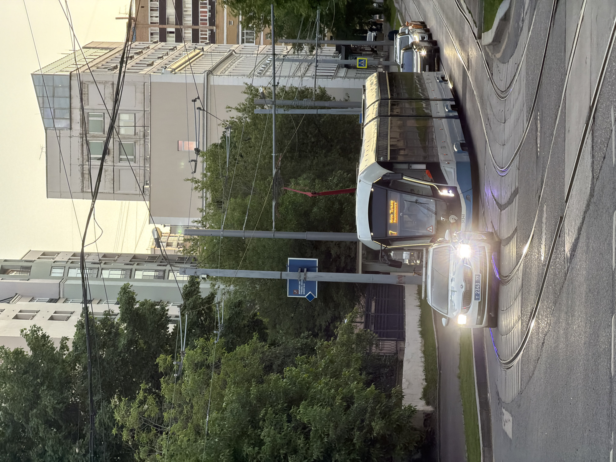 The image shows a tram and a car on a street in Moscow, Russia. The tram is number 7 and displays the Cyrillic inscription "Метро "Бульвар Рокоссовского"." The license plate on the car is T 2343 PP. Buildings and trees line the street. Tram lines run along the road. The light suggests it might be late afternoon or early evening.