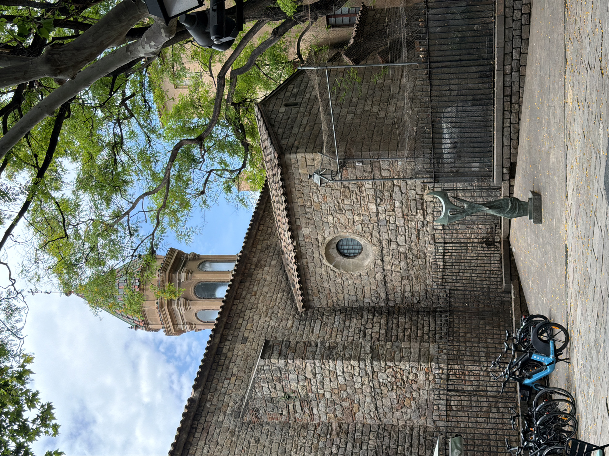 A daytime view captures a historic stone building and its bell tower in Barcelona, Spain, partially obscured by lush green trees. The building is constructed from rough-hewn stone blocks, featuring a terracotta-tiled roof and a circular oculus window. Behind this section, a distinctive bell tower rises, characterized by its ornate, light-colored stone and arched windows, topped with a multi-colored, possibly tiled dome or spire.
In the foreground, a modern, abstract dark metal sculpture stands on a paved surface, which is scattered with small yellow leaves or petals. Adjacent to the sculpture, several bicycles are parked, including a prominent blue one with "BIRD" visible on its frame, indicating a bike-sharing service. A black metal fence separates this paved area from the stone facade of the building.
Overhead, verdant trees with thick branches and bright green foliage extend across the upper and left portions of the frame, contrasting against a clear blue sky dotted with white clouds. The natural lighting suggests mid-morning or afternoon. The scene blends historical architecture with contemporary urban elements, reflecting the character of Barcelona. No people are visible.