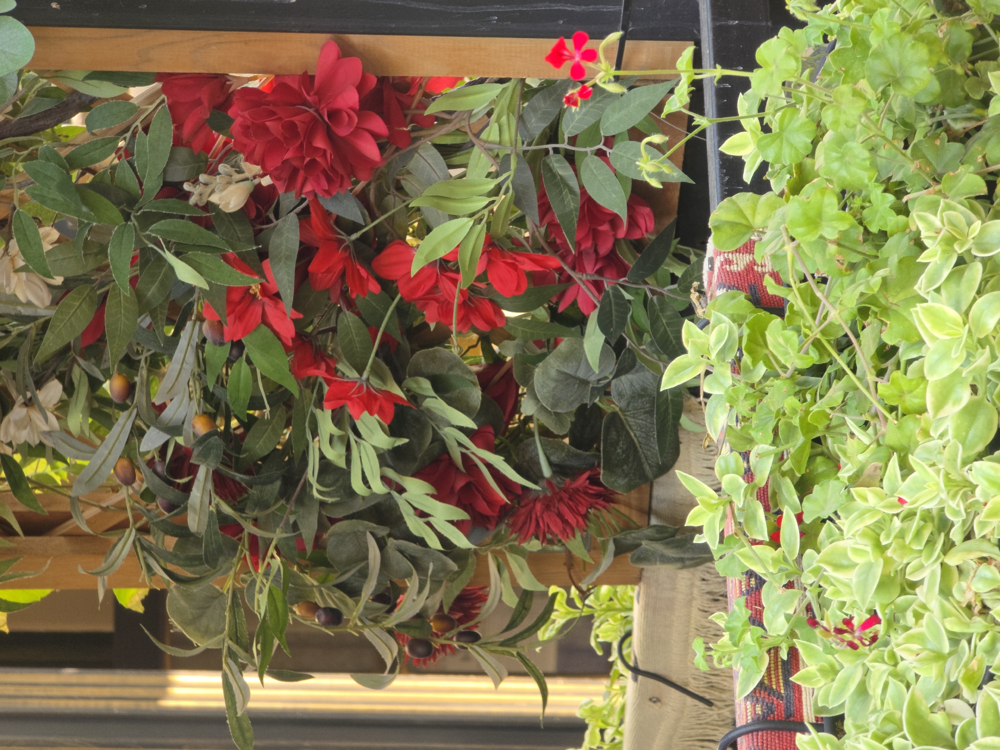 The image shows a close-up of an arrangement of artificial plants, including a faux olive branch with small, dark berries, interspersed with red artificial flowers. The plants are situated within a wooden structure, possibly part of a decorative display or urban greenery. In the foreground, there is a vibrant green ground cover plant. Behind the foliage, a woven fabric with a red and black patterned band is visible. The background is blurred, showing vertical elements that could be part of a building or fence. The lighting suggests an outdoor setting, with sunlight casting shadows. The scene appears to be an exterior decoration in Rostov-on-Don, Russia, likely in a public space or commercial establishment designed to enhance the aesthetic appeal of the environment.