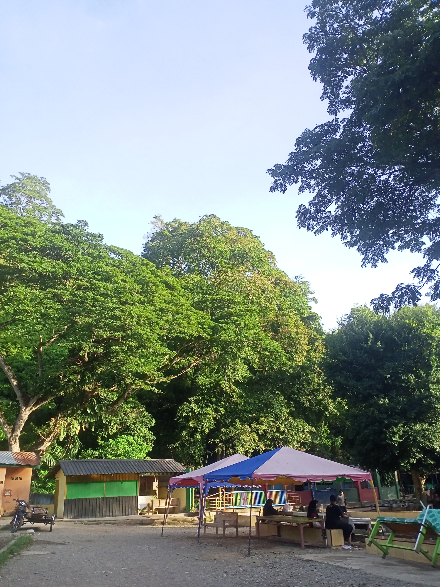 The image depicts an outdoor scene with lush green trees dominating the upper half. In the foreground, there are several small structures that appear to be shops or food stalls with colorful awnings. A blue and pink tent provides shade over a seating area with wooden tables and benches, where several people are seated. To the left, a motorcycle with a small trailer is parked near a building. The ground is covered with gravel. The sky is clear and bright. The scene suggests a relaxed outdoor gathering or a market in a tropical environment. The location is identified as Geundring, Indonesia.