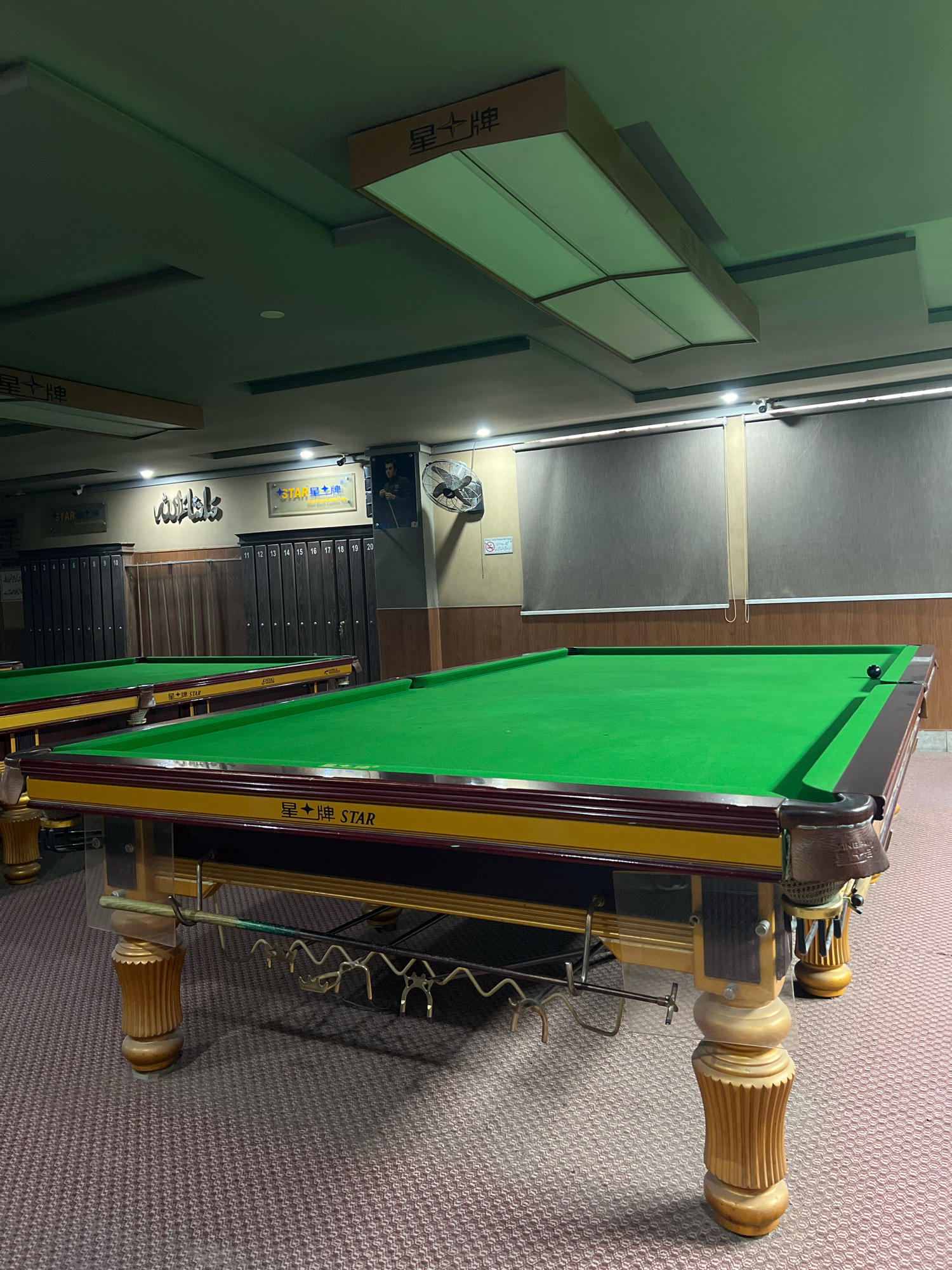 The image depicts the interior of a snooker club in Attock, Pakistan. Two snooker tables with green felt and ornate wooden legs are prominently featured. The table in the foreground is closer to the viewer and is more detailed, showing a dark red and gold trim, along with branding that reads "星牌 STAR". A single black snooker ball is visible on the table's surface. In the background, a series of dark wooden lockers, numbered 11 through 20, are visible. Above the lockers, a framed picture of a man in a suit holding a snooker cue is displayed, next to a wall-mounted fan. Two signs with Chinese characters and the word "STAR" are also present on the wall. The ceiling is a light green color, with rectangular light fixtures and what appear to be acoustic panels. The floor is covered with a patterned carpet in shades of maroon and brown. The overall lighting is dim, with a greenish tint. The scene is static, with no people actively playing or interacting.