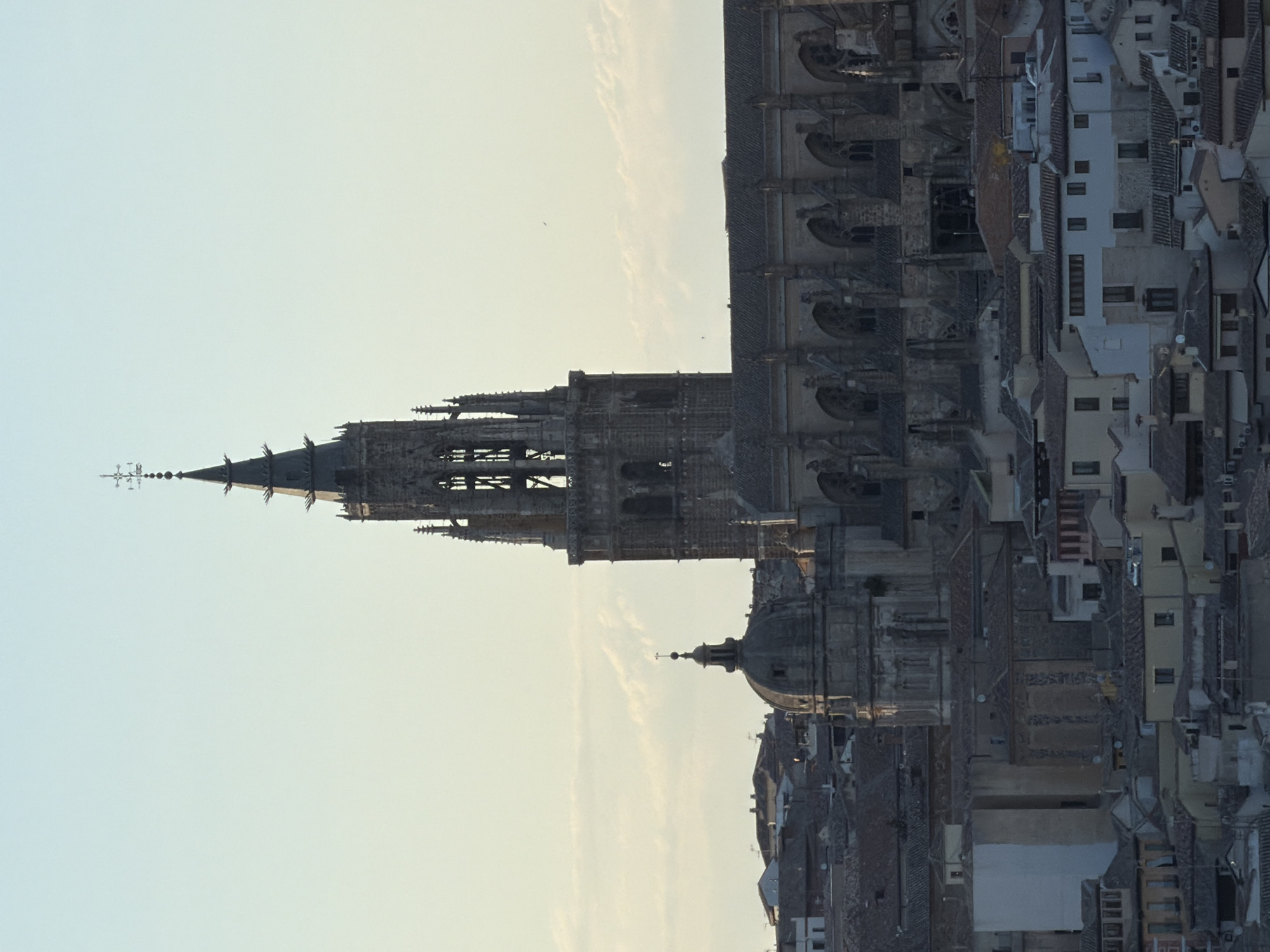 The image displays a panoramic view of the city of Toledo, Spain, dominated by the magnificent Toledo Cathedral. The cathedral's imposing Gothic spire, adorned with a weather vane, pierces the pale blue sky. Below the spire, the intricately detailed facade of the cathedral is visible, featuring a rounded dome to its left. The cathedral is surrounded by a dense urban landscape of terracotta-tiled rooftops and a mosaic of buildings, showcasing a mix of architectural styles. The setting sun casts a warm, soft light on the scene, highlighting the textures and forms of the ancient city. The sky is clear with a few wispy clouds scattered across it. There are no people or animals visible in the image.