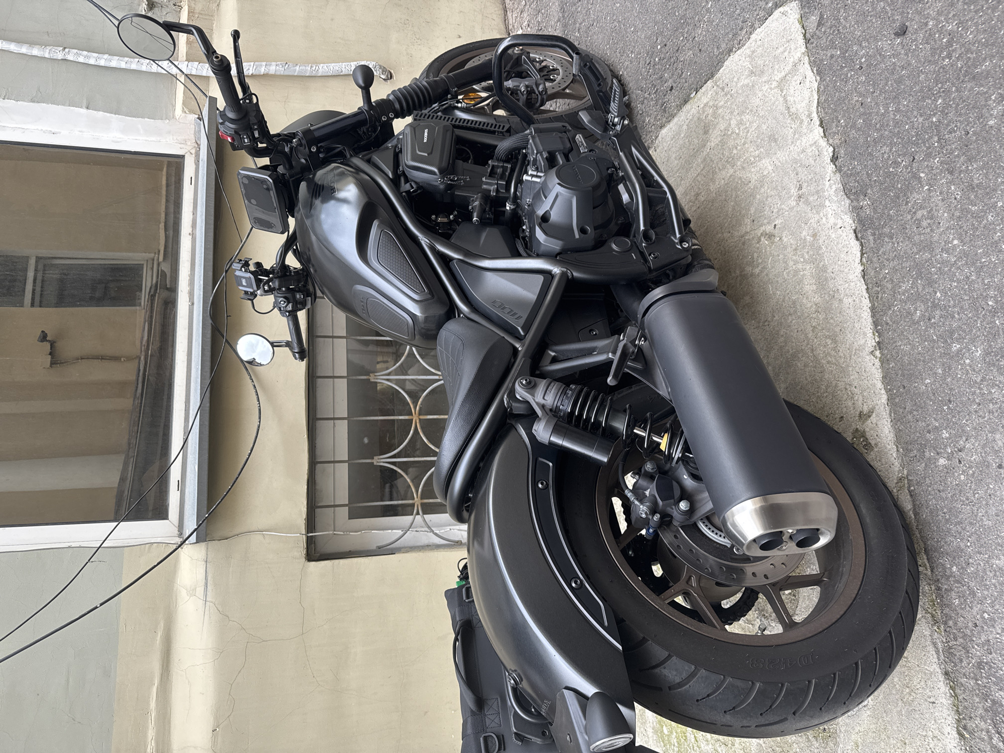 A matte black Honda motorcycle is parked on a concrete slab next to an asphalt sidewalk. The motorcycle is positioned at an angle, facing away from the viewer and slightly to the right. It is a cruiser-style motorcycle with a black leather seat, a black exhaust pipe with a metallic tip, and black handlebars with mirrors and controls. A black saddlebag is attached to the rear left side of the motorcycle.
The motorcycle is parked in front of a light yellow building with a window that has a metal grate. The building appears to be in an urban environment, possibly a courtyard or alleyway, with visible wiring and cracks in the building's stucco exterior. The overall impression is one of a quiet, somewhat aged urban setting. There are no people visible in the image. The lighting suggests it is daytime, with diffuse light suggesting an overcast sky or shade.