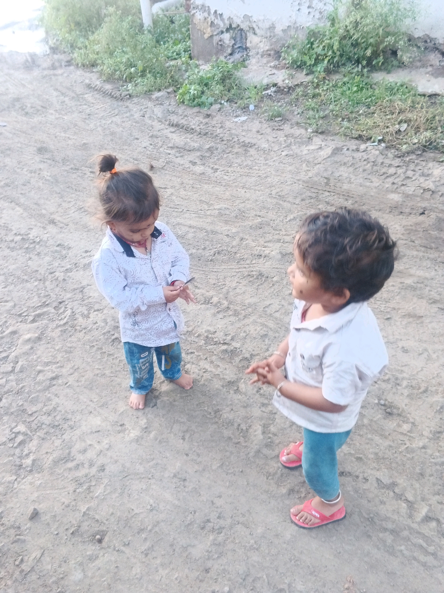 In Rajoda, India, two young children stand on a dirt path. A toddler with dark hair pulled into a small bun wears a light-colored, patterned long-sleeved shirt and blue jeans. They are barefoot and holding a small dark object in their hands. To their right, another child with dark, curly hair wears a light-colored short-sleeved shirt and blue leggings, also with pink flip-flops. This child has their hands clasped together. The ground is composed of loose dirt and pebbles, with tire tracks visible in the distance. Green vegetation grows along a rough, plastered wall behind the children.