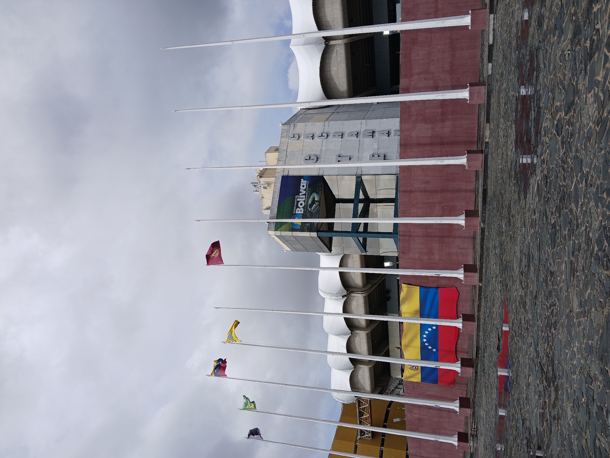 The image shows a view of the Cachamay Stadium in Ciudad Guayana, Venezuela, under an overcast sky. Seven flagpoles are prominently visible, each displaying a different flag. The closest flag is that of Venezuela, recognizable by its horizontal bands of yellow, blue, and red, with a semicircle of stars on the blue band. The other flags are smaller and less distinct. The stadium's facade is a combination of textured maroon paneling and light-colored architectural elements. The ground is paved with dark, irregular stones. A sign with the word "Bolivar" and other smaller text is visible on the stadium's structure. The scene appears deserted.