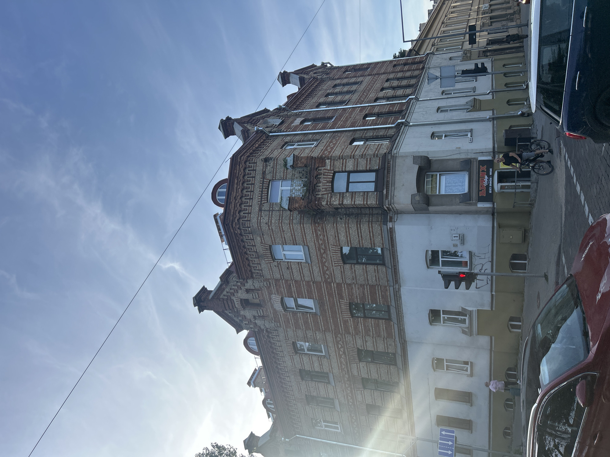 The image depicts a street corner in Vilnius, Lithuania, with a red-brick building occupying the foreground. The building has multiple stories, a distinctive mansard roof with arched windows, and intricate brickwork detailing. A sun flare is visible on the left side of the image, casting a bright light on the building.
In the street, several cars are parked or in motion. A red car is in the lower left of the frame, and a dark blue car is in the lower right. A cyclist is riding a bicycle at the intersection, heading away from the viewer. A pedestrian, a woman wearing a pink shirt and dark pants, is walking on the sidewalk carrying luggage. A traffic light is illuminated red.
A sign for "B-BONX Vintage" is visible on the building's ground floor, indicating a shop. The sky above is blue with scattered clouds, and overhead wires are present. The architecture suggests an older section of the city.