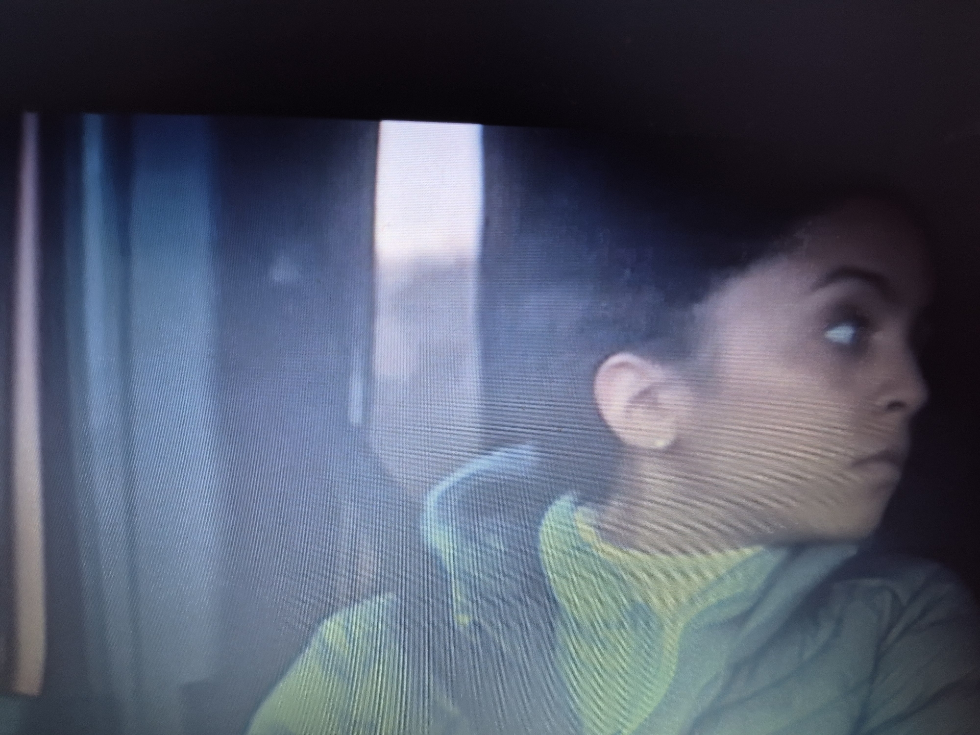 The image shows a close-up of a young woman with dark skin and dark, curly hair, looking to her right. She is wearing a light green hooded jacket over a yellow turtleneck. A small, pearl-like earring is visible in her left ear. The background is dark and out of focus, with vertical bands of light and shadow suggesting an indoor setting, possibly a vehicle or a room with diffused lighting. The texture of the image shows digital artifacts, likely due to a screen capture. There is no discernible indication of the location within Sandwell, United Kingdom.
