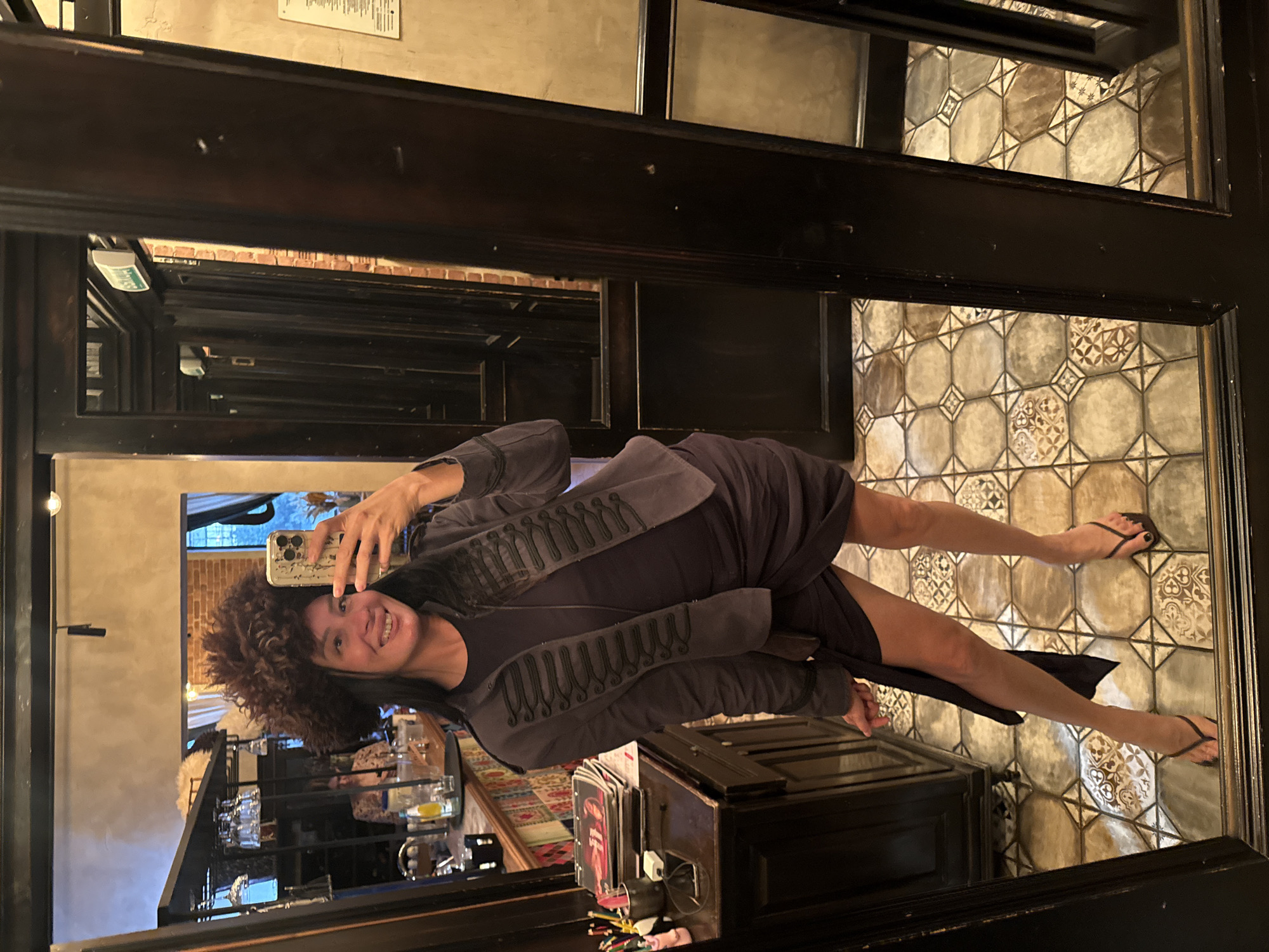A woman is taking a selfie in a mirror, wearing a large, fluffy brown hat and a dark purple dress with a matching military-style jacket. She is in what appears to be a bar or restaurant with a dark wood interior. The floor is tiled with an intricate geometric pattern. In the background, reflected in the mirror, are shelves with glassware, a bar counter, and other patrons. The overall impression is of a stylish and playful pose in an atmospheric setting. The scene is likely indoors, possibly in a dimly lit establishment.