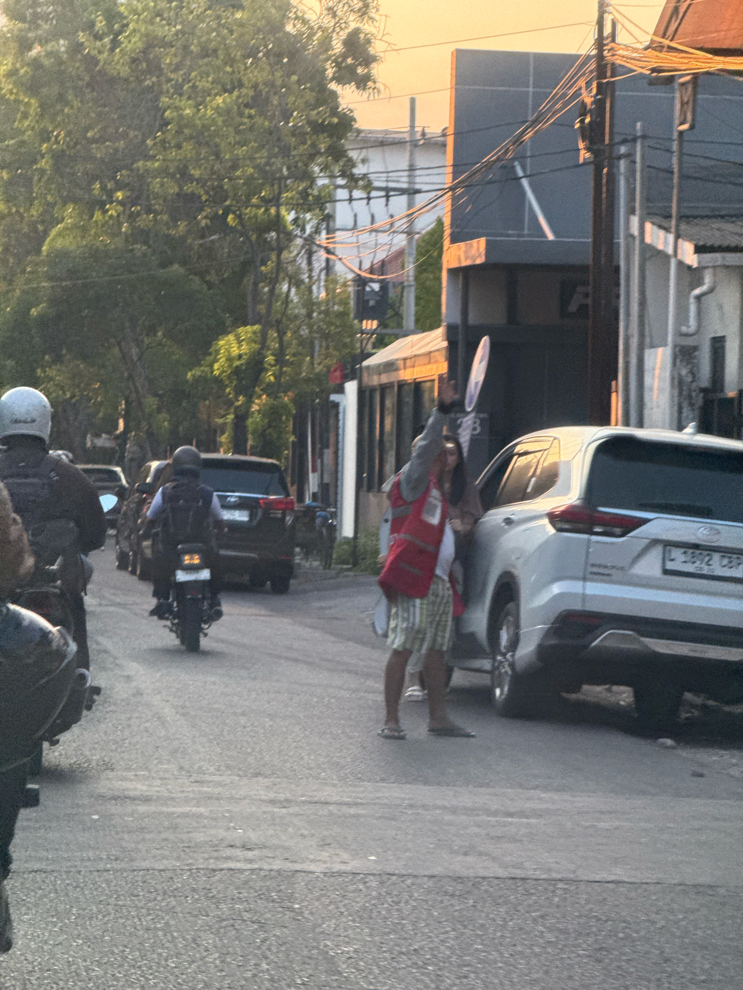 The image captures a street scene in Surabaya, Indonesia, likely during late afternoon or early evening, given the warm, soft lighting. In the foreground, a person wearing a white helmet is seated on a motorcycle, facing away from the camera. Further down the road, another person on a motorcycle, also wearing a helmet, is visible. A dark SUV is positioned in the middle ground, with its rear visible. To the right of the SUV, a person in a red vest and checkered shorts is seen holding a round, white object aloft, seemingly directing traffic or signaling. Another individual, a woman, is standing beside the SUV with her hand on the open door. The environment includes trees lining the street, buildings in the background, and overhead power lines. The road surface appears to be asphalt.
