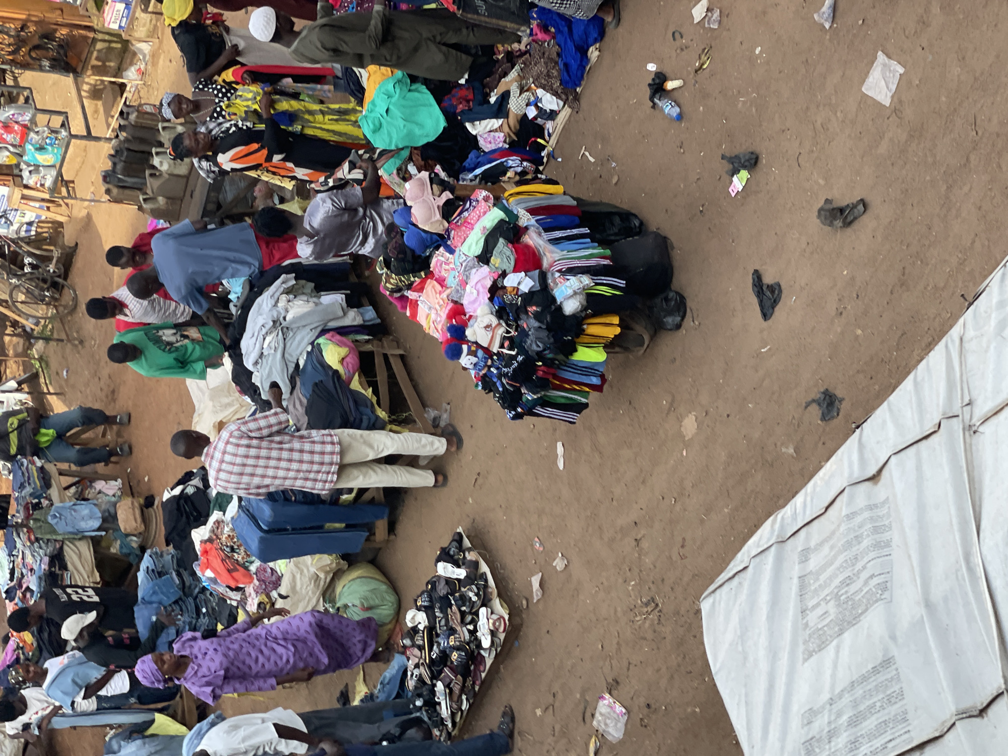 The image shows an outdoor market scene in an unidentified city. A number of people are clustered around piles of clothing, likely for sale. They sit on chairs or low benches while looking at the clothes. There are piles of shoes also offered for sale. The ground is bare dirt, scattered with bits of trash and debris. A large white tent-like structure occupies the right-hand side of the image.