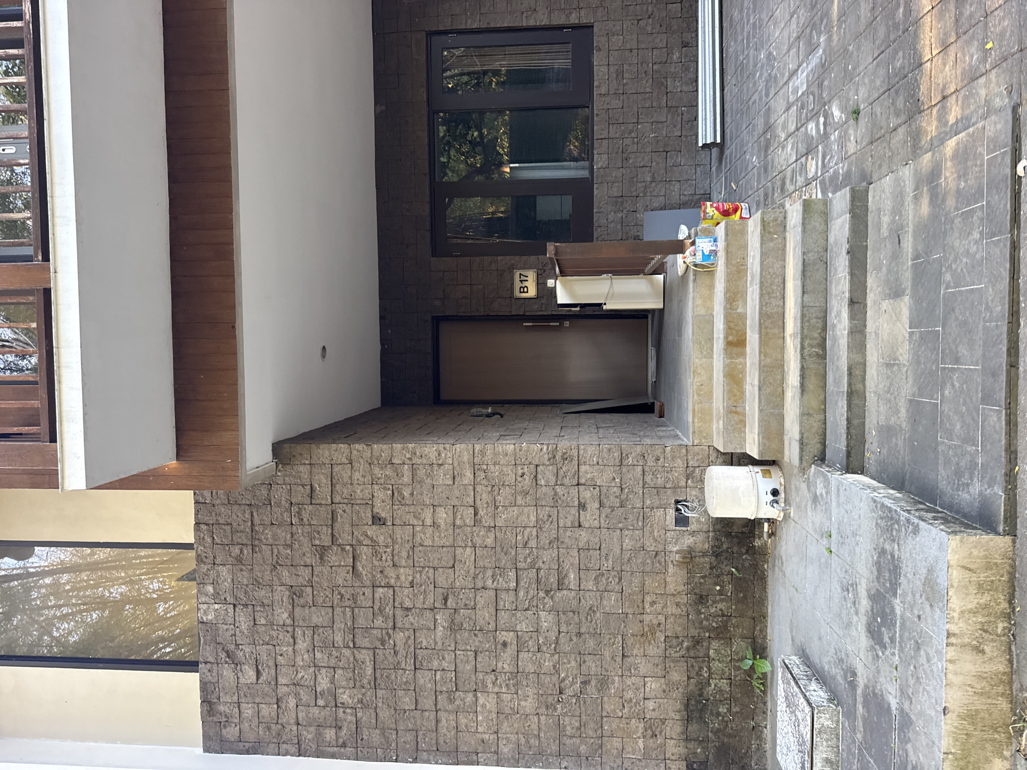 In Kuta Selatan, Indonesia, a front view of a multi-story building shows a facade of natural stone tiles. A narrow concrete pathway leads to a set of stone steps. To the left of the steps, a white cylindrical water heater sits on a concrete slab. A wooden door is visible behind the steps, with a sign reading "B17" above it. A large brown-framed window is to the right of the door. The building has a wooden balcony on the upper level. The ground is paved with gray stones. Sunlight casts shadows across the scene.
