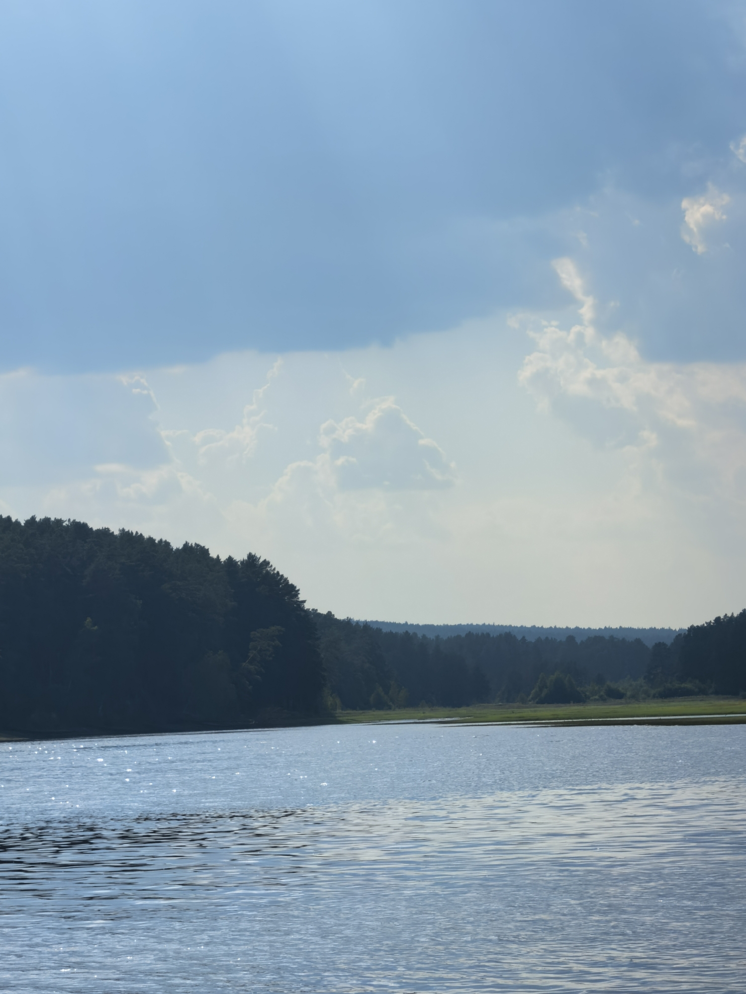 This media file depicts a serene lake scene in Sysert, Russia. The primary subjects are the vast expanse of water, a densely forested shoreline, and a sky filled with dramatic clouds. The image captures a tranquil natural setting, with no people or man-made objects immediately visible.
The activity taking place is simply the stillness of nature. The water's surface is gently rippled, reflecting the sky and sunlight, creating a shimmering effect. The shoreline is a dark, silhouetted line of evergreen trees, suggesting a temperate climate. In the distance, the treeline continues, with a strip of lighter green grass or meadow visible between the water and the distant forest.
Notable details include the time of day, which appears to be late afternoon or early evening. The sunlight catches the water, creating bright highlights, and the shadows are lengthening. The weather is dynamic; while the sky is blue, it is interspersed with large, fluffy white clouds that suggest some atmospheric activity, perhaps indicating a partly cloudy day. The location is identified as Sysert, Russia, which adds a geographical context to the landscape. The overall emotion evoked by the image is one of peace, solitude, and the quiet beauty of the natural world. There is no visible text in the media file.
