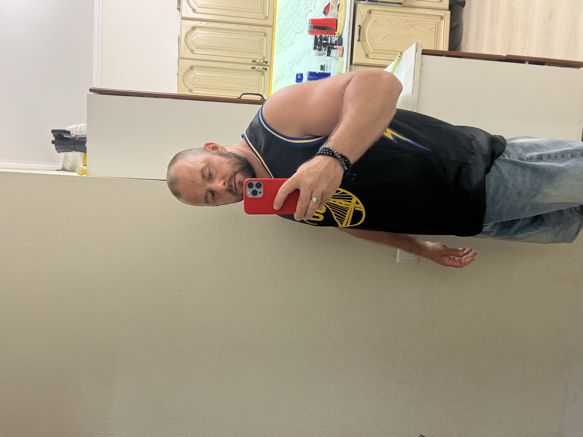 A bald, Caucasian man in his late 30s or early 40s with a beard, wearing a black Golden State Warriors jersey and denim shorts, takes a selfie in what appears to be a kitchen. He is holding a red smartphone in his right hand, which is raised to eye level. The man is standing in front of a textured, light-colored wall. To his right, a white kitchen cabinet is visible, topped with various items, including a black coffee maker and a yellow object. Further to the right, light-colored kitchen cabinets with ornate detailing are mounted on the wall, and a white countertop displays several bottles and containers, including a red electric kettle. The floor is light-colored wood. The overall environment suggests a residential kitchen. The image does not contain any specific visual cues that would allow for precise identification of the location within the city of Sochi, Russia.