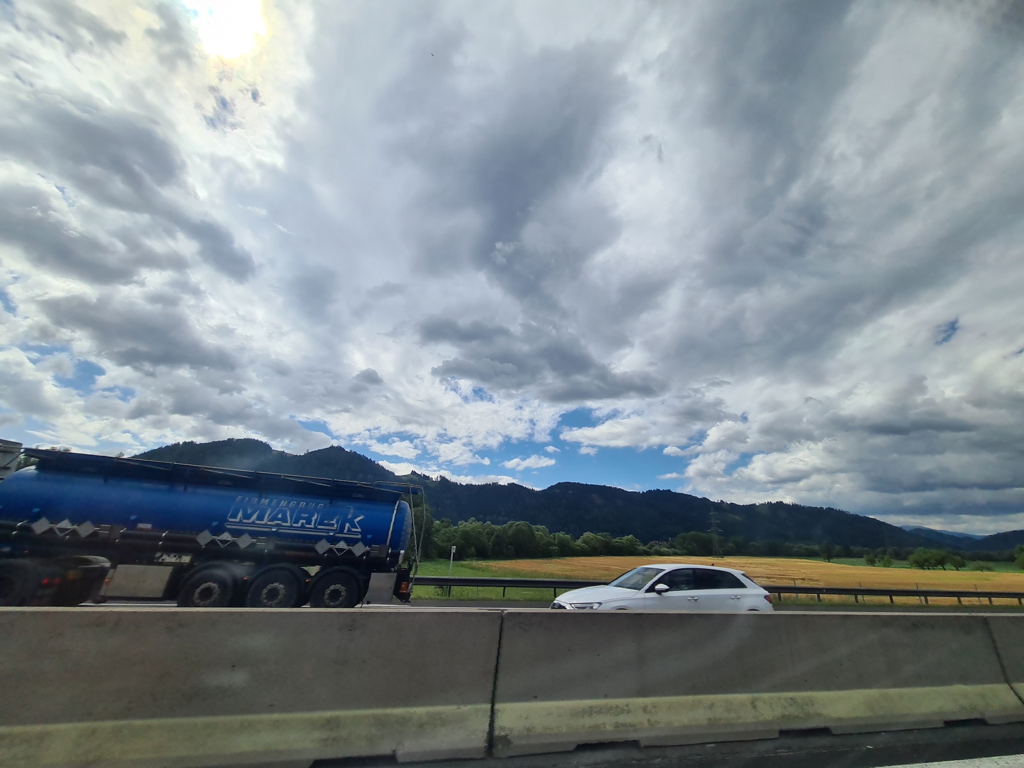 The image captures a highway scene in the vicinity of St. Stefan ob Leoben, Austria, under a dramatic sky with a mix of sun and clouds. In the foreground, a concrete barrier runs across the bottom of the frame. To the left, a blue tanker truck with "MAREK" and "FIRMENGROUP" written on its side is in motion. It has multiple axles and is likely carrying a liquid. To the right of the truck, a white sedan is also traveling on the highway. Behind the vehicles, a yellow field stretches towards a dark green, tree-covered mountainside. The sky above is filled with heavy, gray clouds interspersed with patches of blue sky and bright sunlight peeking through. The overall impression is one of travel on a clear but potentially inclement day in a rural Austrian landscape.