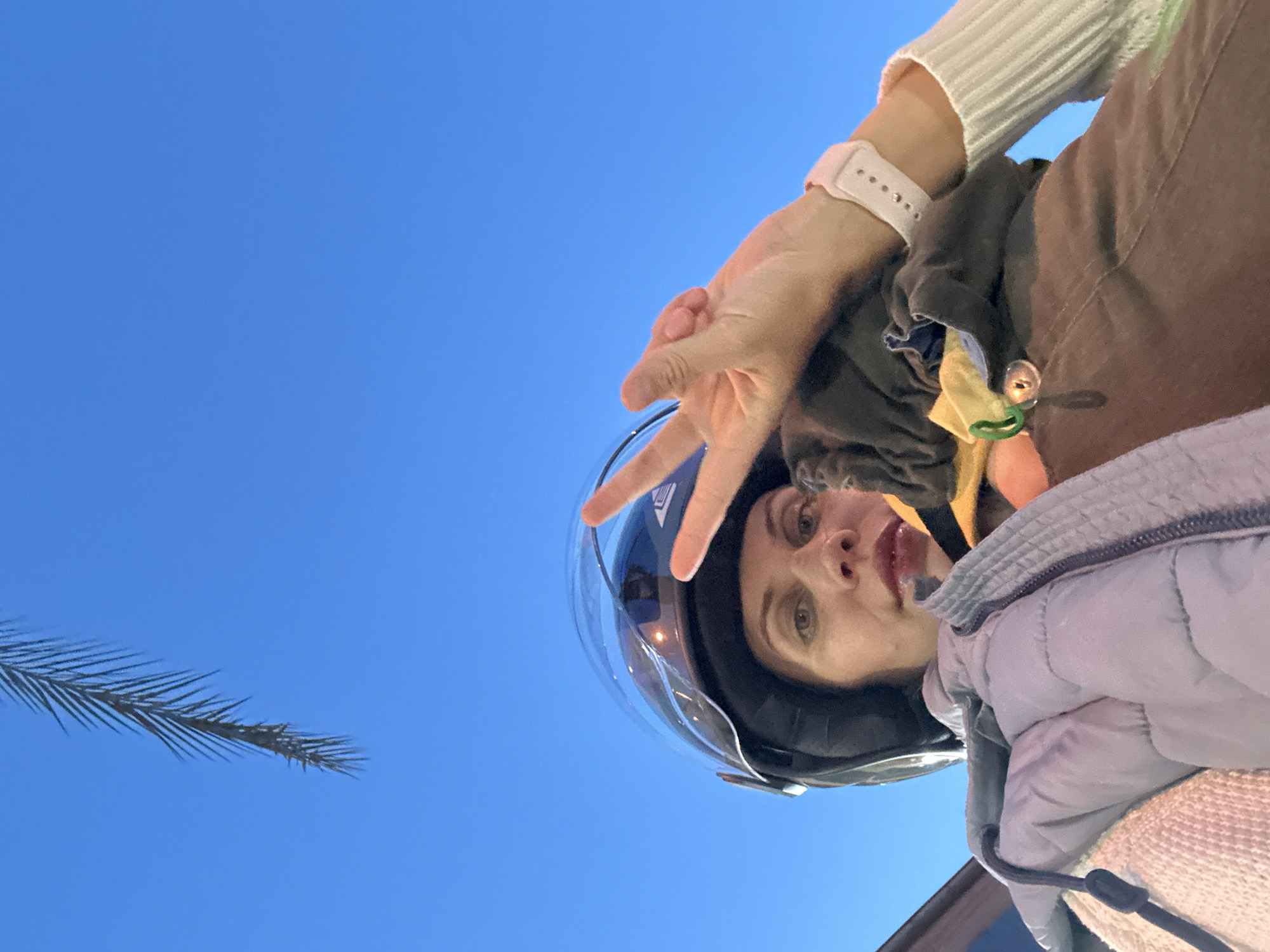 A woman wearing a helmet is pictured in a selfie against a bright blue sky. She is flashing a peace sign with her right hand. The helmet has a clear visor. She is wearing a light-colored sweater and a puffy jacket. A palm frond is visible in the lower left corner. The location is Fethiye, Turkey.