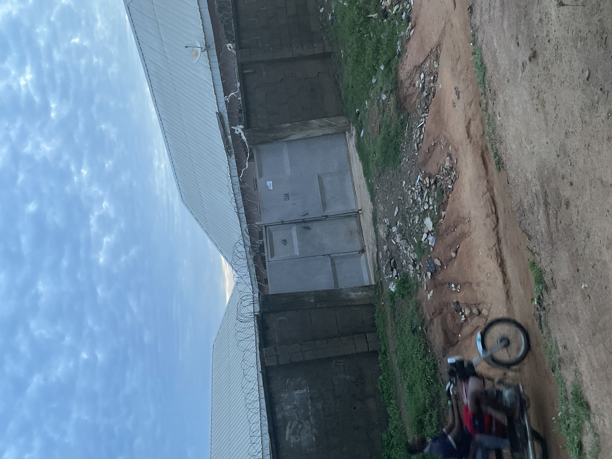 The image depicts a cloudy sky over a compound with a metal roof and a brick wall topped with barbed wire. Two large, gray metal gates are visible in the wall. A person is riding a motorcycle, blurred by motion, on a dirt road in the foreground. The environment appears to be a somewhat unkempt urban or semi-urban area, with some debris scattered around. The scene is likely located in Jalingo, Nigeria, a city known for its typical architecture and road conditions.