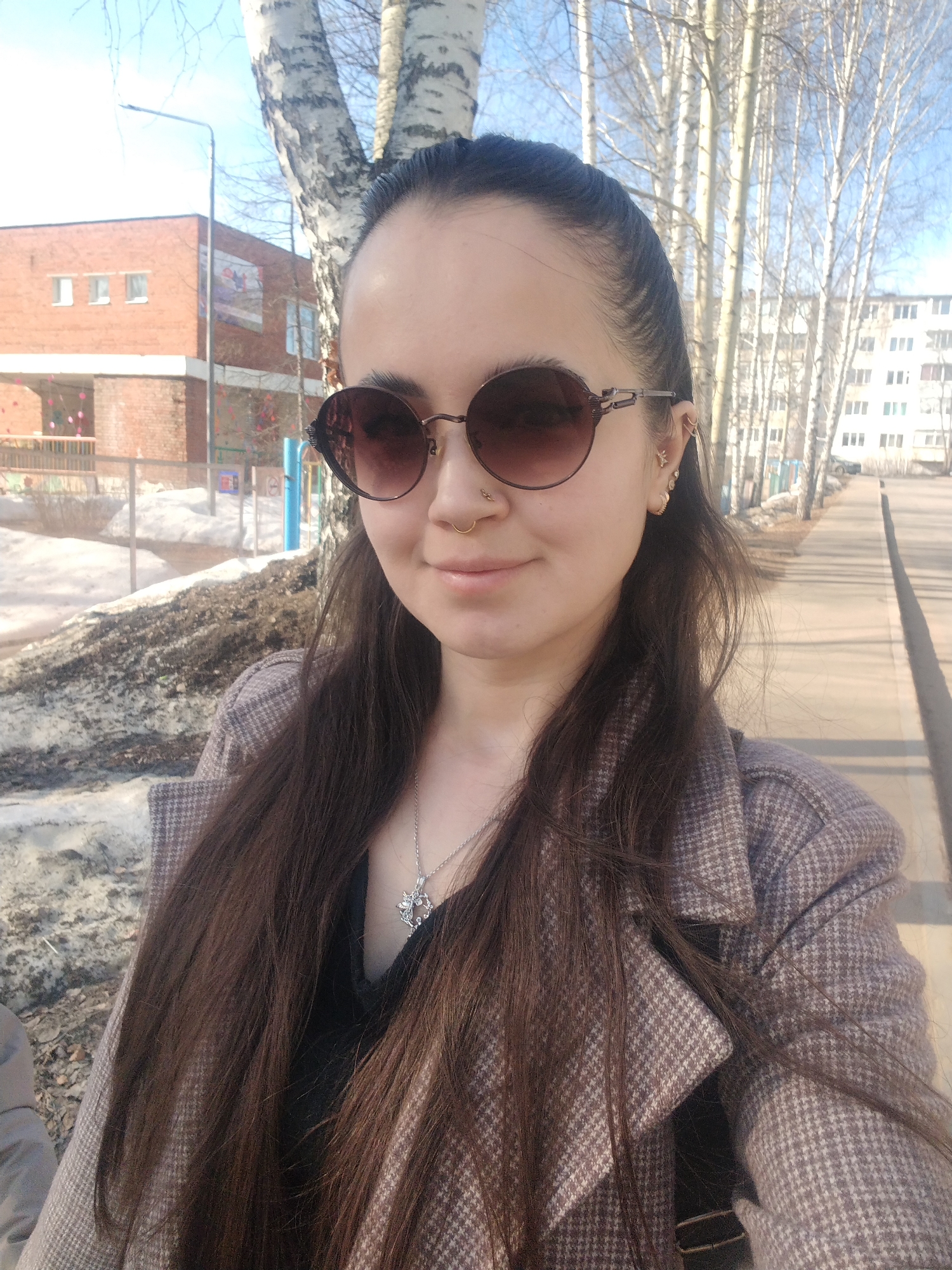 A woman in a brown patterned coat and round sunglasses takes a selfie outdoors. She has long brown hair pulled back from her face and is wearing a necklace. In the background are birch trees, a sidewalk, and buildings characteristic of urban landscapes. The presence of snow suggests early spring. A closer examination indicates the building on the left is a brick structure, possibly a school or kindergarten, and the taller building on the right appears to be a residential apartment complex. The scene suggests a location in Noviy, Russia.