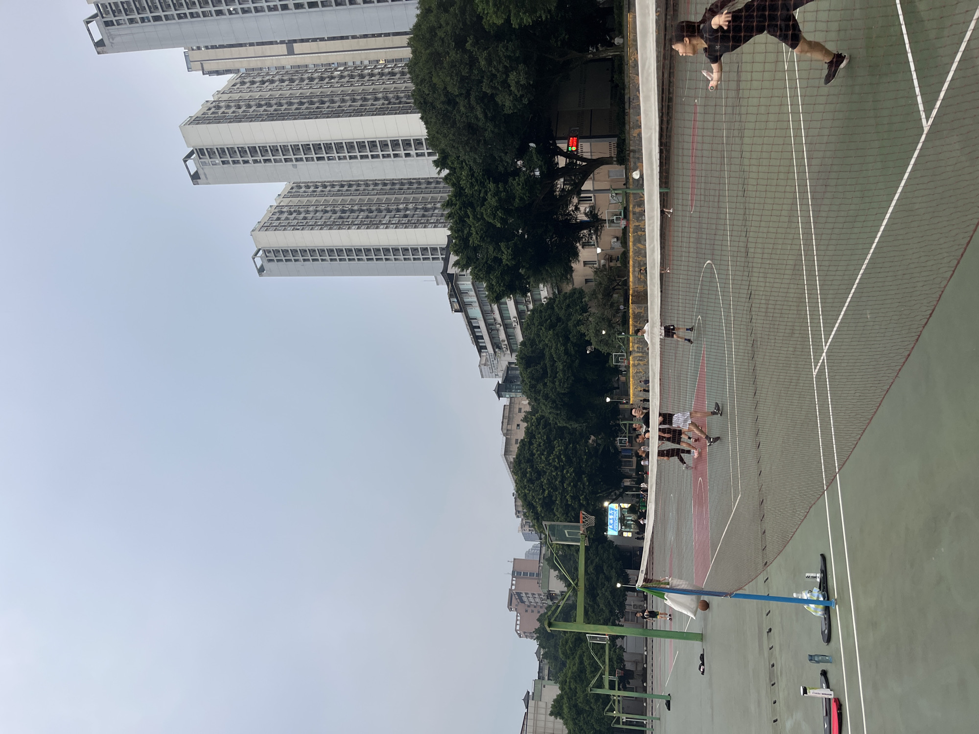 The image depicts an outdoor recreational area, likely a park or sports complex, in Yuzhong District, China. In the foreground, a badminton net divides the court, and two individuals are engaged in a game. One person, dressed in black athletic wear, is actively playing on the right side of the net, in mid-swing with a racket. On the left side of the net, another person in white and dark shorts is positioned on the court. In the background, several basketball hoops are visible. Further back, a group of people are seen near a basketball court, and some are walking. Tall residential buildings and dense foliage, including trees, form the backdrop against a hazy sky. The overall scene suggests an urban setting with integrated recreational facilities.