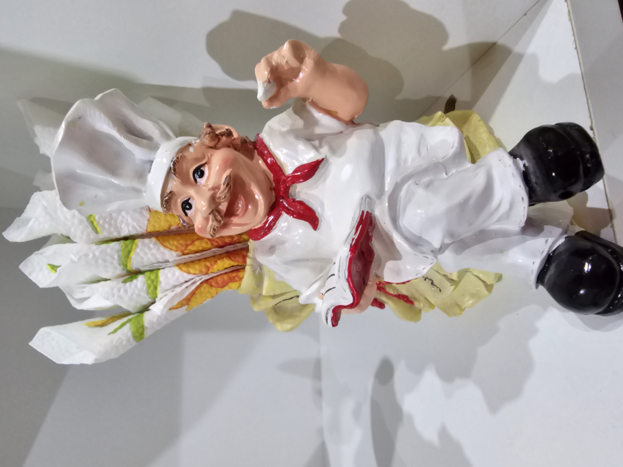 This is a close-up, eye-level, indoor shot of a ceramic figurine of a chef holding a small book in his left hand and a piece of food in his right. The chef is wearing a white chef's hat, a white shirt with a red bandana, and white pants with black shoes. The figurine is seated on a yellow base that resembles a flower, and behind the chef is a bouquet of paper napkins with a floral pattern. The background consists of a white shelf and a wall, with shadows cast on the wall. The figurine is positioned to the right of the frame, with the left side of the image being mostly empty space. The image was taken in Kalinovka, Russia.