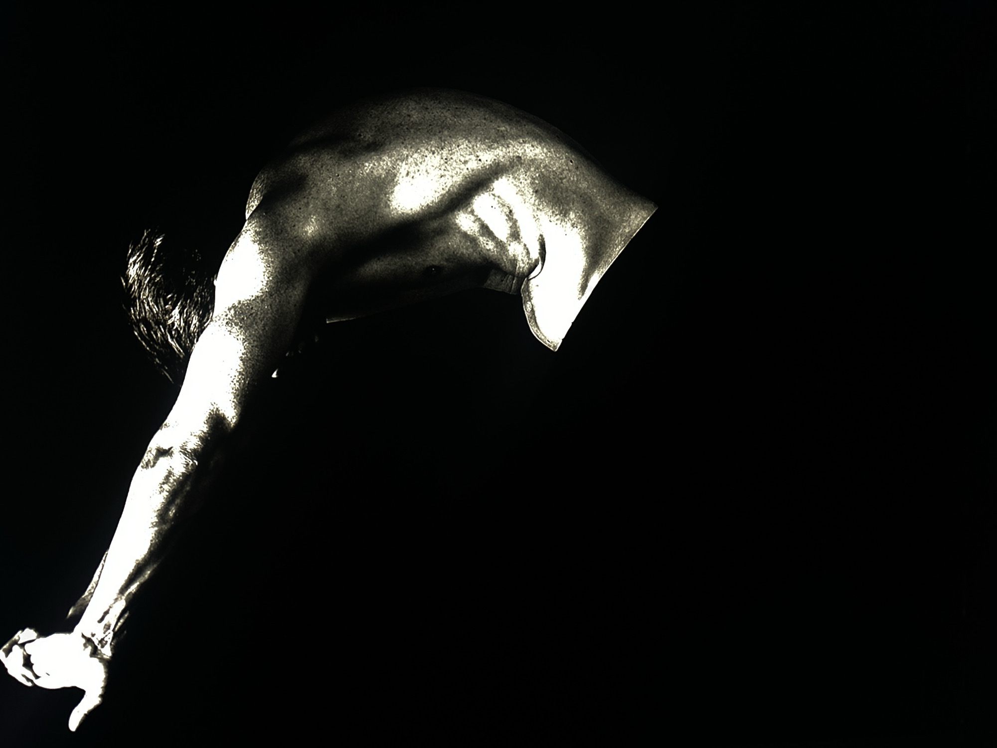 A black and white image shows the upper torso and arm of a man bent over, positioned on the left side of the frame against a completely black background. The man's skin is illuminated, showing muscular definition in his back, shoulder, and arm. His head is bent down with wet-looking hair. The image's perspective and lack of context don't provide any clues about the location, making it impossible to determine any association with Заволжье, Russia.