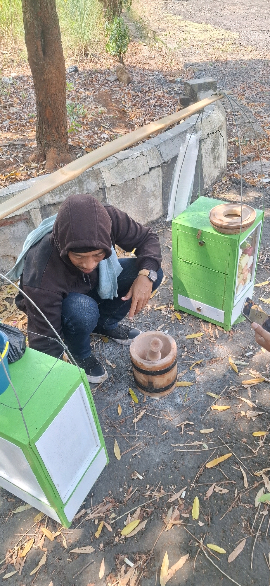 In Cilaku, Indonesia, a man wearing a brown hooded sweatshirt and blue jeans is squatting outdoors. He has a light blue cloth draped over his shoulders and is wearing a watch on his left wrist. In front of him, on the ground, is a wooden mortar with a pestle. To his right is a green wooden food cart with a glass display case filled with cut fruit. The cart has a circular wooden object on top. To his left, another green food cart is partially visible. The ground is covered with dirt, fallen leaves, and twigs. In the background, a tree trunk and concrete structures are present.