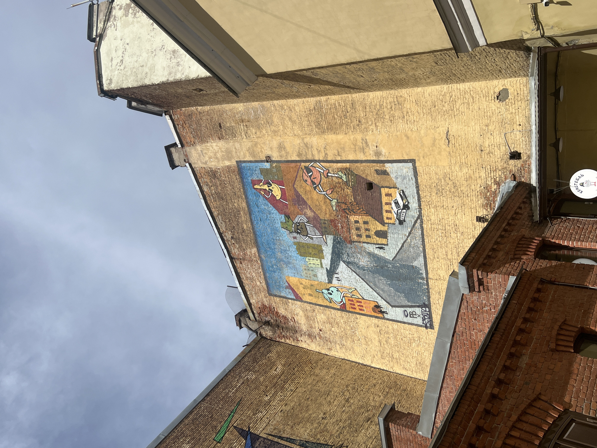 The photograph shows a mural on the side of a brick building in Saint Petersburg, Russia. The mural depicts cartoon-like figures against a cityscape background, featuring bright colors. The wall supporting the mural is textured with visible brickwork and faded yellow paint. Surrounding the building with the mural are adjacent structures made of brick and plaster, with varying architectural details, including windows, roofs, and decorative elements. The sky is visible in the background, exhibiting overcast conditions.