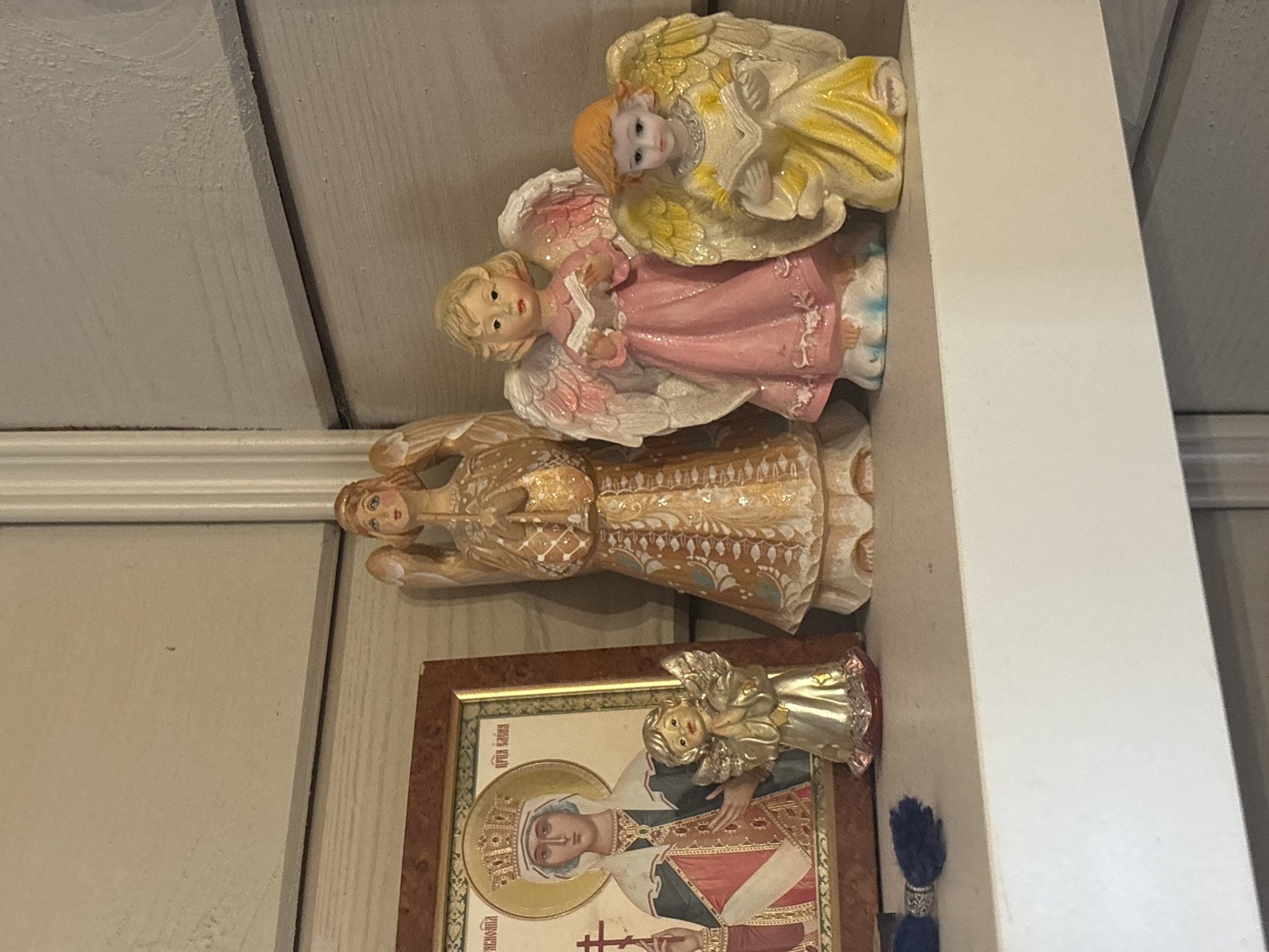 The image displays a shelf in a home at Березка Снт (поварово), Russia. On the shelf, there are several angel figurines and a framed religious icon. The icon depicts a female figure with a crown, holding a cross. One angel figurine is small and gold, with its hands clasped in prayer. To its right, a larger angel figurine stands with its hands also clasped in prayer and wears a patterned golden robe. Further to the right, two more angel figurines are present; one is wearing a pink dress and holding an open book, and the other, smaller figurine, is in a yellow dress and also holding an open book. The background shows a textured wooden wall and a white ledge. There are no people visible in the image.