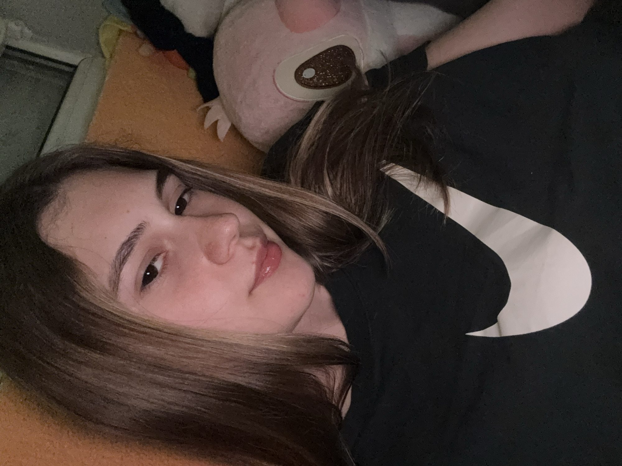 A young woman is captured in a close-up, laid-back shot, likely in an indoor setting such as a bedroom or living room in Panagyurishte, Bulgaria. She is lying on her side, with her head resting, and looks directly at the camera with a neutral expression. Her long, brown hair, with subtle lighter streaks, cascades over her shoulders and part of her face. She has visibly glossy lips.
She is dressed in a black top, which appears to be a t-shirt or hoodie, featuring a large, white Nike swoosh logo prominently displayed on the chest area. The surface she is lying on is an orange or brownish textured material, possibly a blanket or bedspread. Behind her head, a light pink plush toy with distinctive large, glittery brown eyes is visible. The background also includes glimpses of what appears to be a wall or a window frame. The lighting in the image is soft and somewhat dim, creating a relaxed atmosphere, suggesting it might be evening or night, or simply an interior space with subdued lighting. The overall impression is one of casual repose.