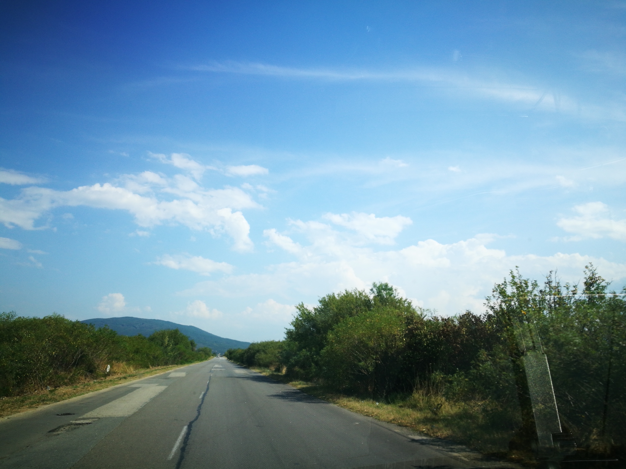 The image depicts a daytime scene of a road winding through a rural landscape. The road is paved and appears to be in moderate condition, with some patching and cracks visible. The sky is a clear blue with scattered white clouds. On either side of the road, there are lush green trees and vegetation. In the distance, a large hill or mountain is visible. The overall impression is one of a scenic drive through a natural environment. The image was taken from a moving vehicle, as evidenced by the slight blur and the perspective. There are no people or animals visible in the frame.