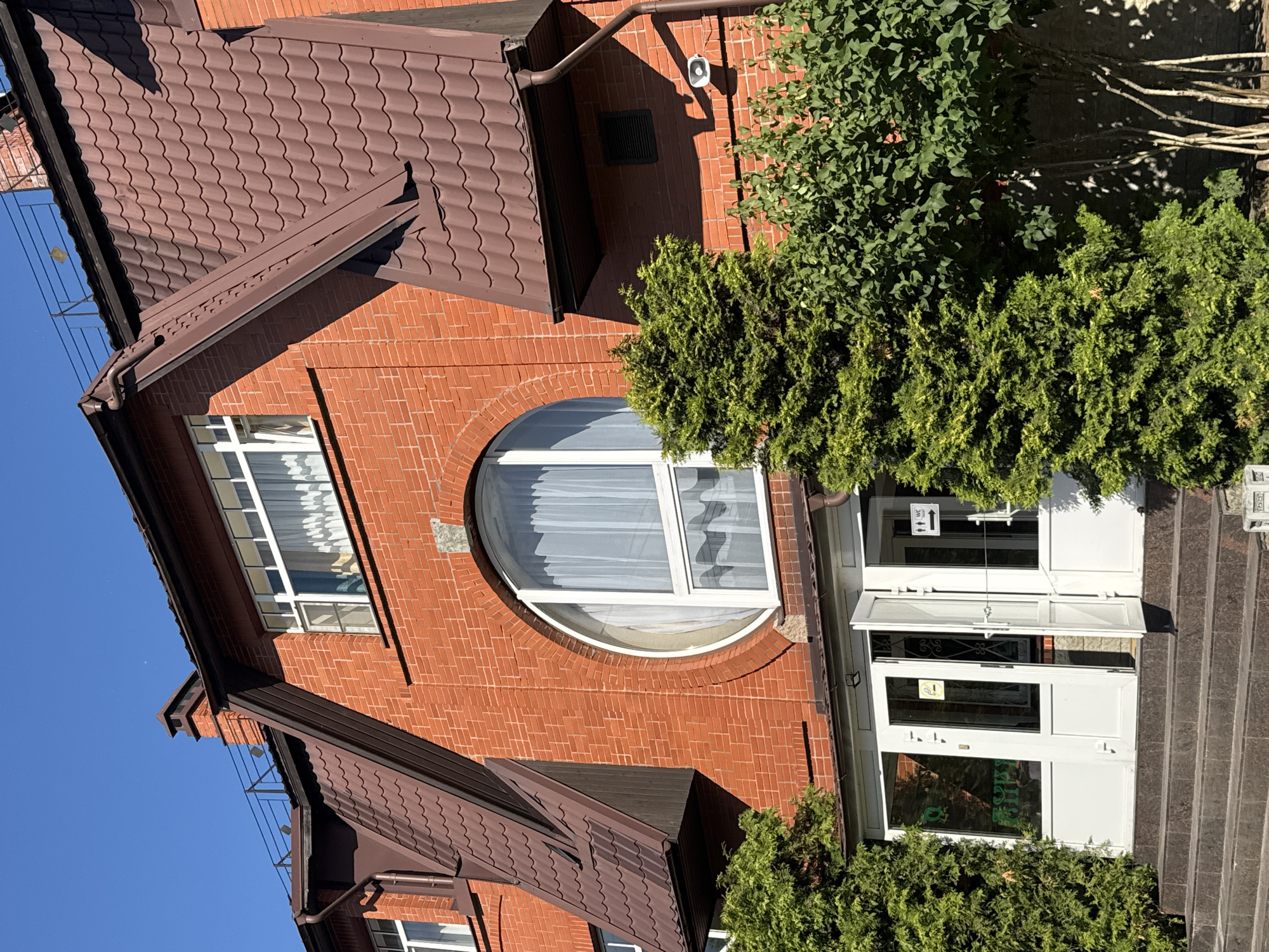 The image shows the exterior of a brick building with a brown tiled roof, set against a clear blue sky. The building features several windows, including a large, semi-circular window on the upper floor with white curtains. There are also double glass doors leading into the building, one of which is propped open. Green bushes and trees are visible around the building, suggesting an outdoor setting. The lighting indicates it is daytime, likely a sunny day. A sign with symbols resembling a male and female figure and the letters "WC" is visible above the entrance. Another sign with yellow and black text is on the lower door. The image is taken from a slightly elevated angle, looking down at the building's facade and entrance. The architectural style suggests a modern or renovated building. The overall scene appears to be a commercial establishment or a public building. There is no visible indication of specific activity or event taking place beyond the static view of the building and its surroundings. The location context of Saint Petersburg, Russia, is provided but not visually evident within the image itself.