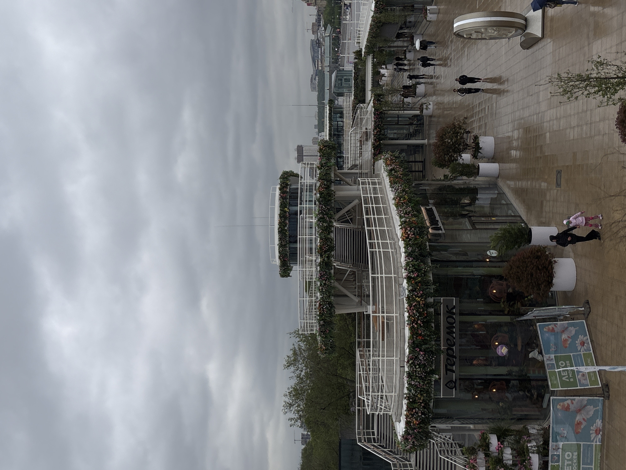 The image shows a section of the Zaryadye Park in Moscow, Russia, on a cloudy day. The view is elevated, looking towards the park's pavilion buildings with white railings and floral decorations. Pedestrians are walking on a brick-paved pathway. The building in the foreground has "Teremok" signage. The background shows the cityscape of Moscow with buildings and communication towers.