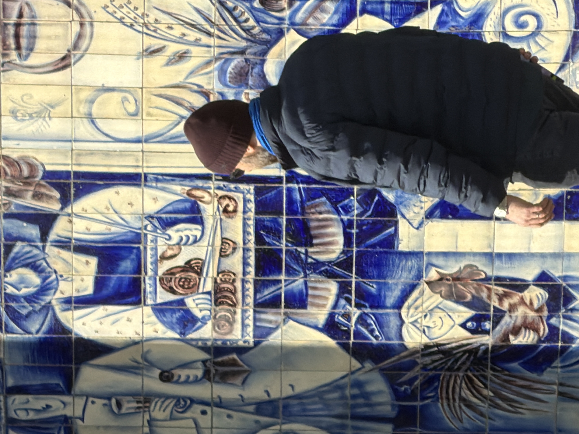 A male individual is observed from behind and slightly to the right, standing approximately one meter from a tiled wall mural within the Chernyshevskaya Metro Station in Saint Petersburg, Russia. The individual wears a dark brown knit beanie, sunglasses, a visible beard, a blue neck gaiter, a dark blue puffer jacket, and dark trousers. A metallic watch is present on his left wrist, and a ring on his left ring finger. His posture indicates he is observing the mural.
The wall is adorned with a large ceramic tile mural executed in shades of blue and cream. The mural depicts stylized human figures engaged in activities and holding various objects, characteristic of Soviet-era art. Key figures include:
- On the left, a tall, slender figure in a light-colored suit holds a stemmed glass. Below this, a smiling figure with long hair holds a rooster and green foliage.
- In the upper center, a figure wearing an apron and a head covering is depicted holding a tray containing multiple circular food items (e.g., pastries or bread) and utensils.
- The mural also incorporates decorative elements such as swirls, floral patterns, and what appear to be musical instruments or traditional implements. The tiles are arranged in a grid, and the scene is uniformly lit, suggesting an indoor environment.