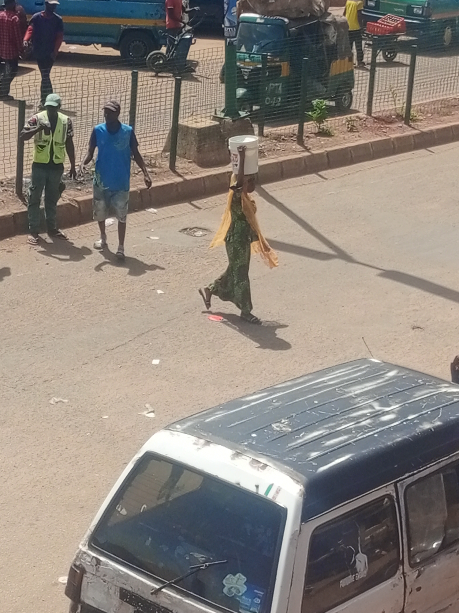 A street scene in Bukuru, Nigeria, depicts a woman walking with a white bucket on her head. She is wearing a green patterned dress and a yellow headscarf. Two men stand on the sidewalk in the background. The man on the left wears a yellow vest and green pants, while the man on the right wears a blue t-shirt and denim shorts. A blue bus is visible in the distance, along with a green auto-rickshaw. The ground is paved with asphalt, and a wire fence separates the sidewalk from the street. The weather appears to be sunny, with shadows cast on the ground.