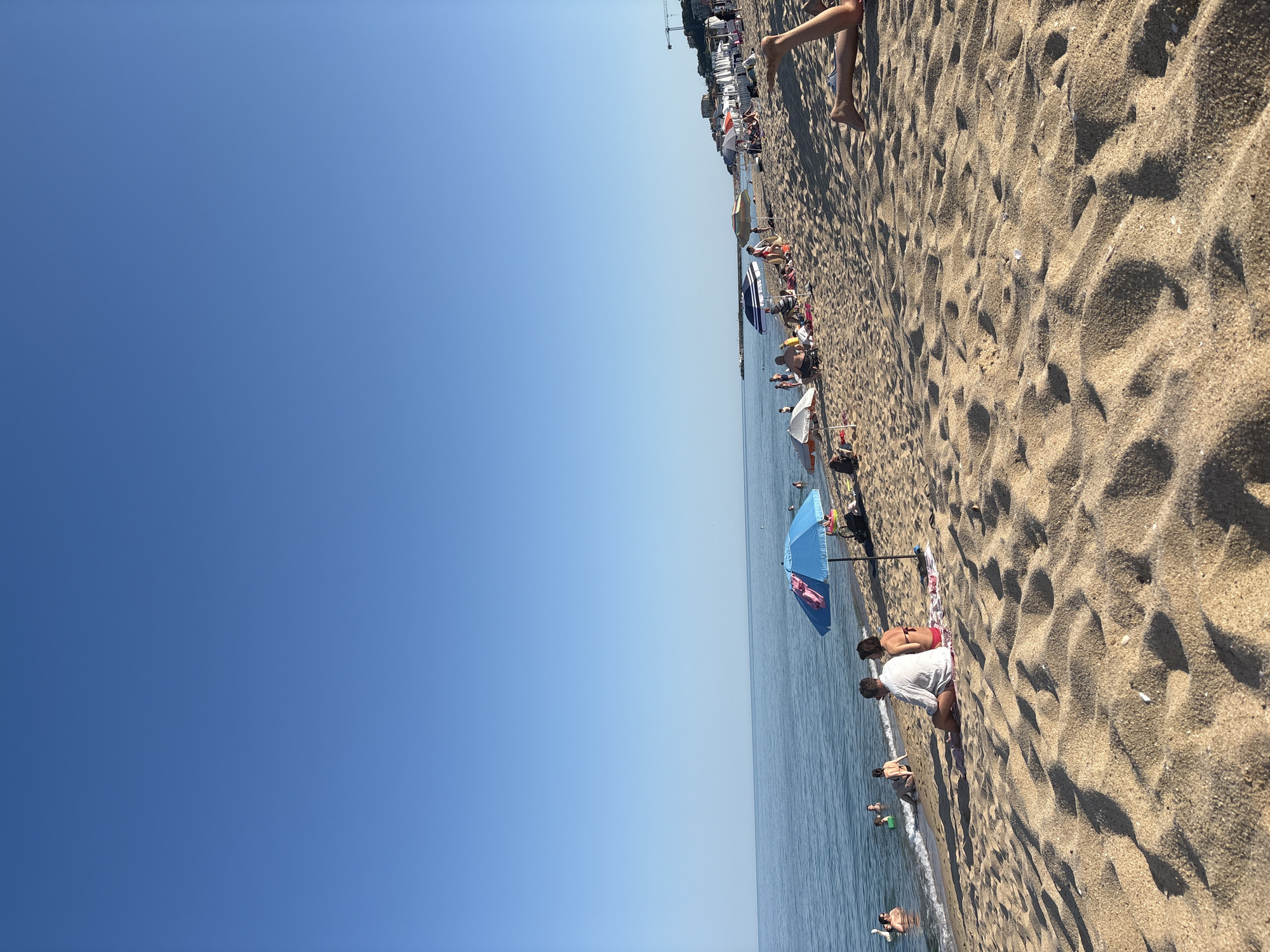 A wide shot captures a bustling beach on a clear, sunny day. The sand is a light tan, marked with the footprints and patterns of many visitors. A vast expanse of calm, blue water stretches to the horizon, meeting a cloudless sky of the same serene blue.
Numerous people are scattered across the beach, engaged in various summer activities. Some are sunbathing on towels or chairs, while others are wading or swimming in the gentle waves. Beach umbrellas, predominantly in shades of blue and white, provide pops of color and shade. One particularly vibrant blue umbrella stands out near the water's edge.
In the foreground, the texture of the sand is clearly visible, with the shadows of people and objects casting long shapes. Further back, a line of beach chairs and umbrellas suggests a more organized section of the beach, with buildings and perhaps a crane visible in the distance along the coastline.
The overall atmosphere is one of leisure and enjoyment, typical of a summer beach day. The bright sunlight indicates it is likely mid-day, and the clear sky suggests fair weather. The scene is one of a popular vacation spot, with many people seeking to enjoy the sun, sand, and sea.
