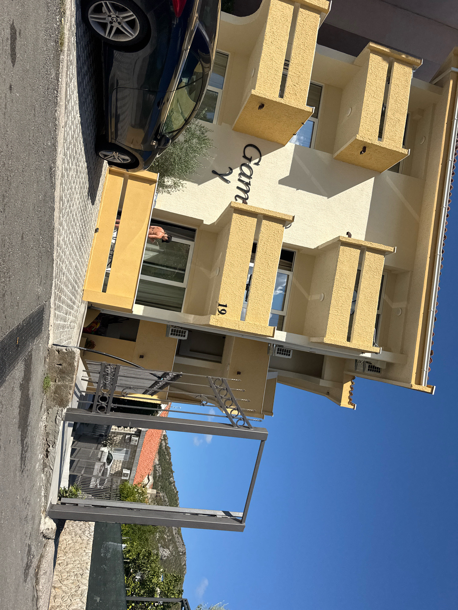 A dark-colored BMW is parked on a patterned cobblestone driveway next to a pale yellow building. The building has multiple balconies with decorative stone accents and a sign that reads "Gammy." A person is visible through a window on the second floor of the building. The scene appears to be in Bečići, Montenegro, with a view of a mountainous landscape and the blue sky in the background. The image is shot from a slightly low angle, with the building dominating the right side of the frame.