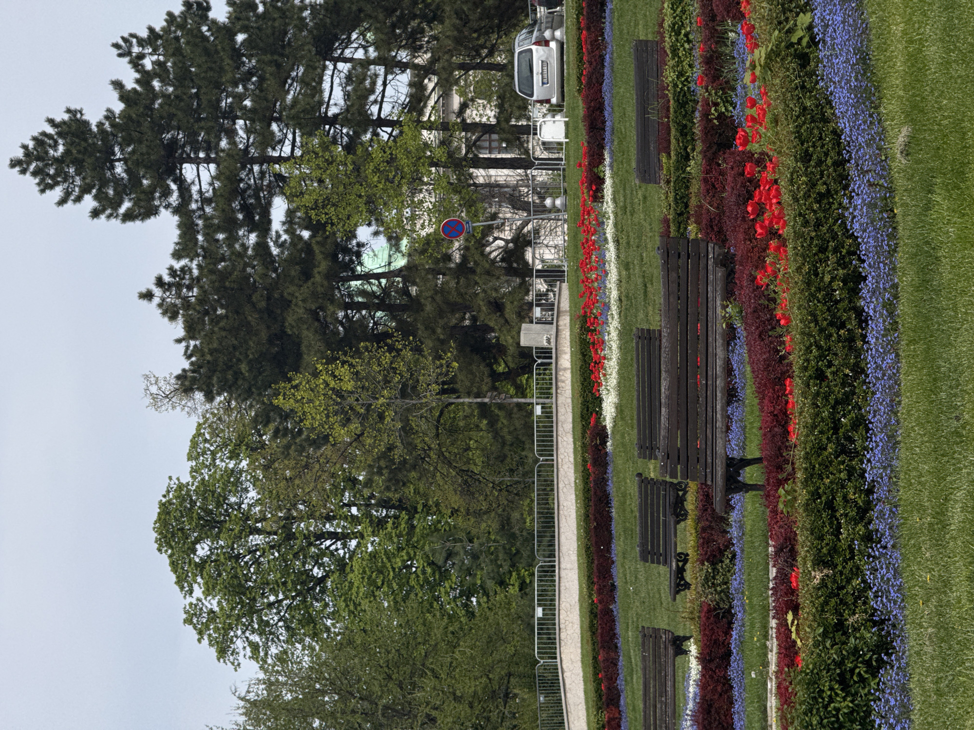 The image depicts a park scene in Belgrade, Serbia. Rows of flowers in various colors, including red, white, and blue, are arranged neatly along a grassy area. Park benches are situated adjacent to the flower beds. In the background, trees and buildings are visible, with a silver vehicle parked near one of the structures. A pedestrian path runs alongside the flower beds and benches, partially lined with a metal fence. A road sign is also visible. The sky is overcast.