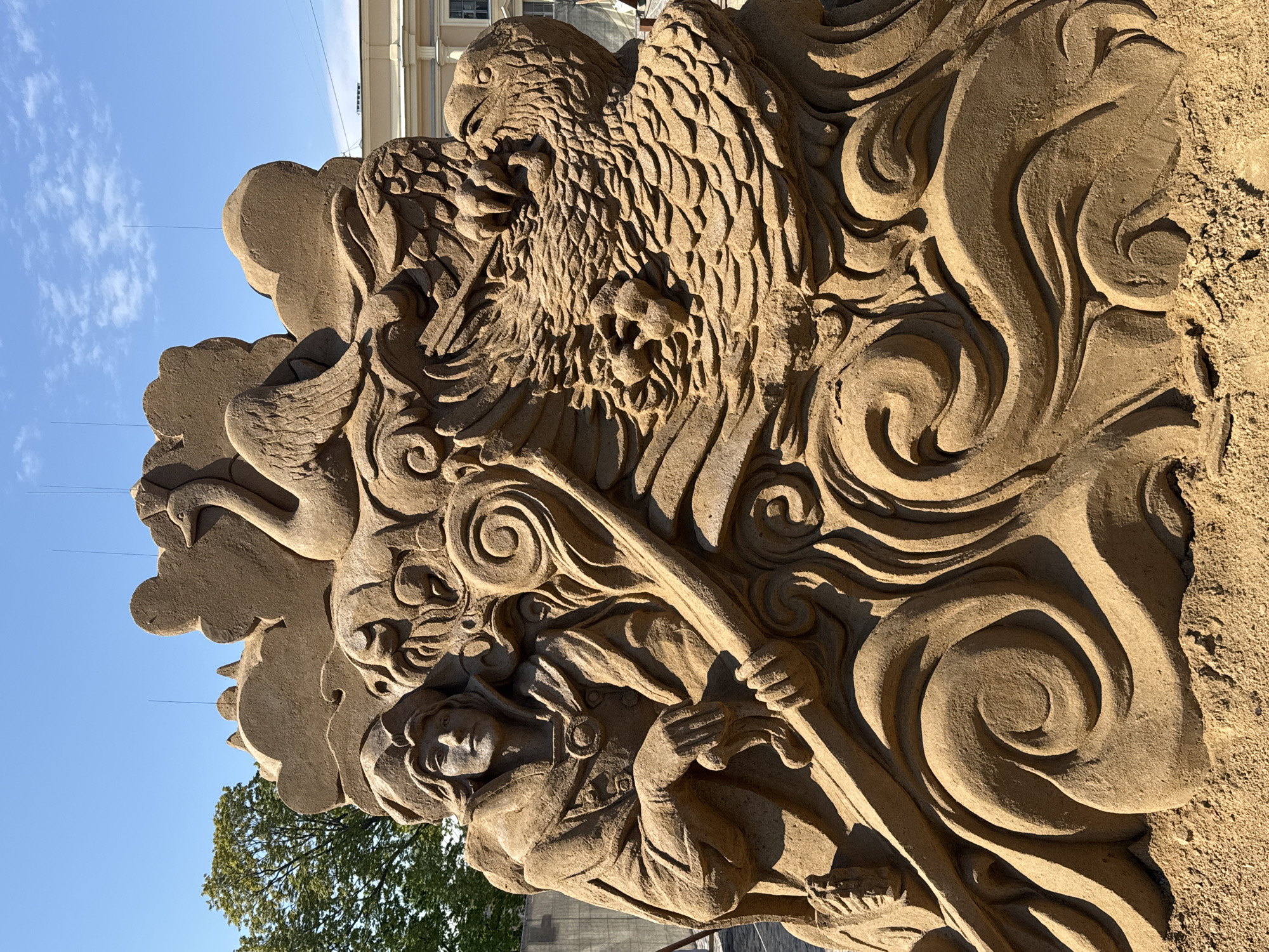 This is a detailed sand sculpture, likely part of an art installation or festival. The sculpture features an elaborate design with an eagle at the top, waves, a person holding a pole, and a swan. The backdrop shows a clear blue sky and the top of a building. The sculpture may be located in Saint Petersburg, Russia.