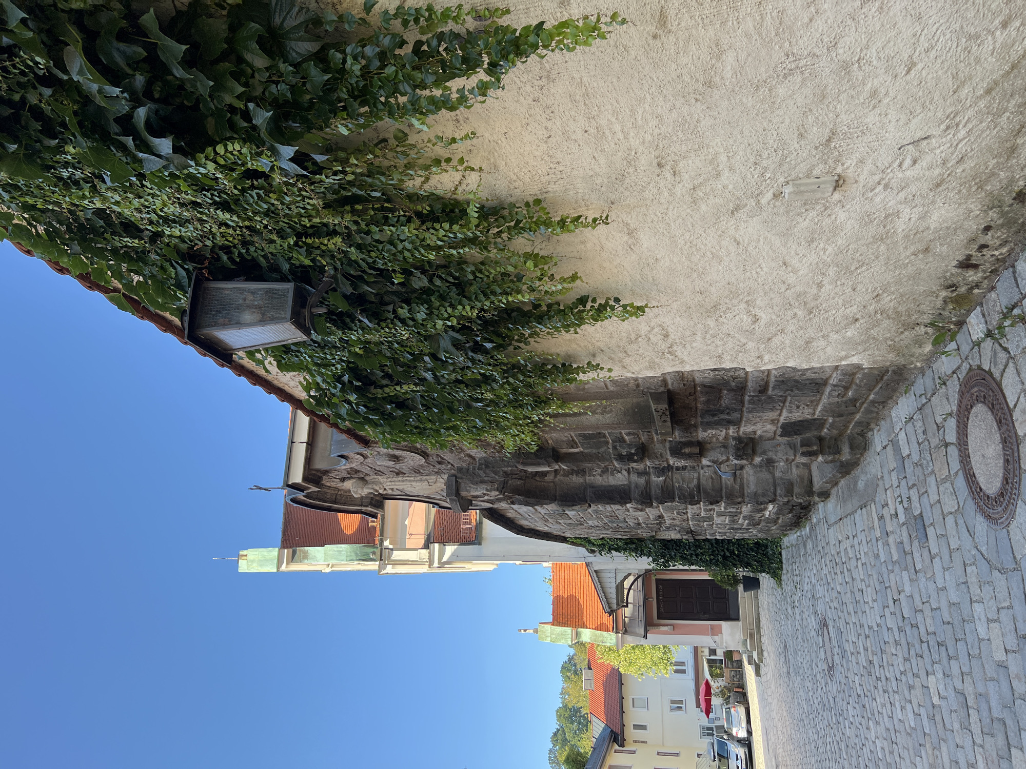This image depicts a cobblestone street in Immenstadt im Allgäu, Germany, leading towards several buildings. In the foreground, a weathered wall covered in lush green ivy dominates the right side of the frame, with an antique lantern affixed to it. To the left, the street curves slightly, revealing buildings with red-tiled roofs and pale facades. One building features a prominent arched doorway and a small green spire. The sky above is a clear, bright blue. The scene appears tranquil, with no people visible. The architecture suggests an older European town.