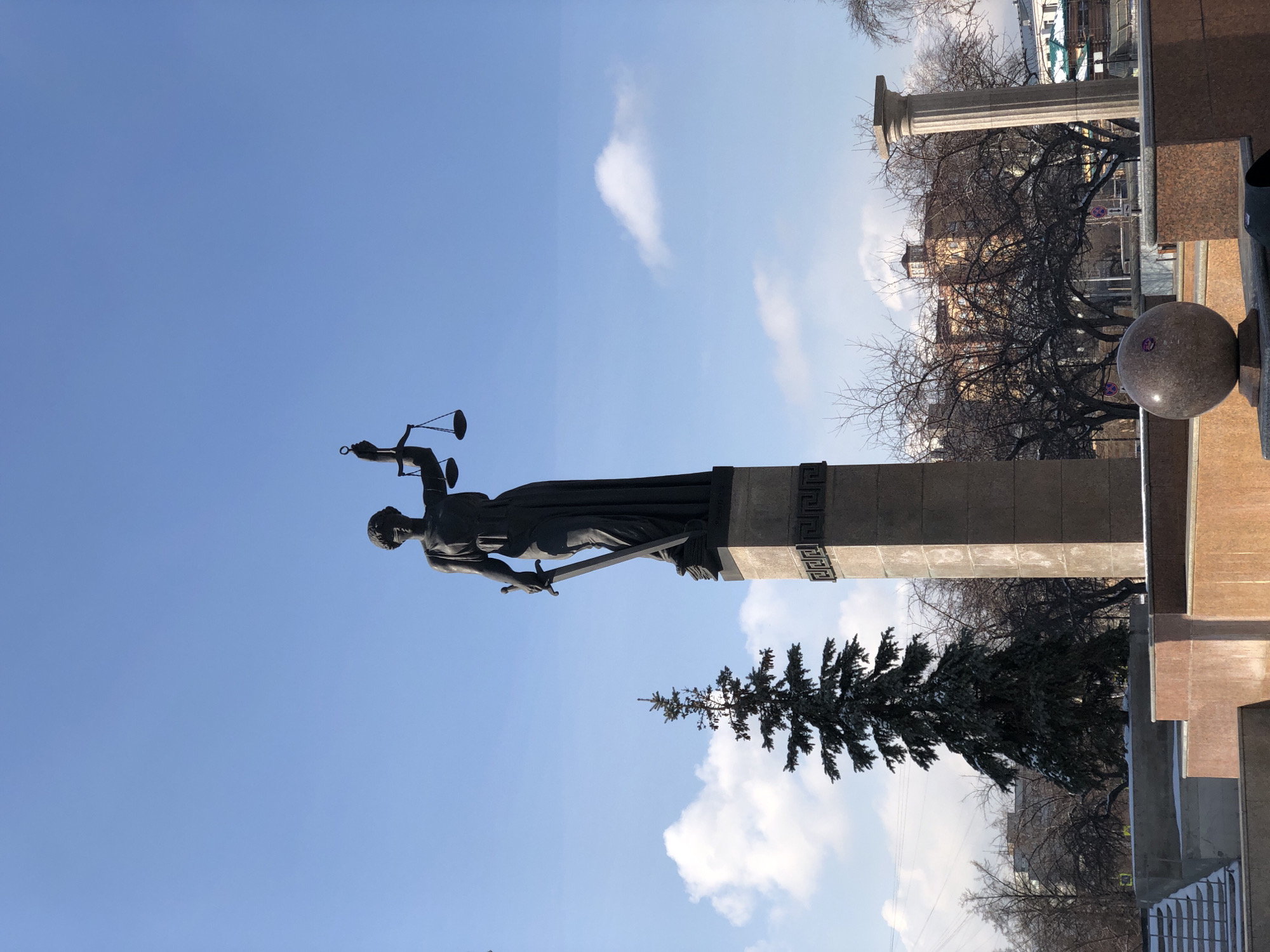 The image displays a monumental statue of an allegorical figure, likely Lady Justice, against a bright, clear blue sky dotted with scattered white clouds. The dark-colored statue, possibly bronze, stands prominently on a tall, rectangular stone pedestal. The figure holds the scales of justice in her raised left hand and a sword, pointed downwards, in her right hand. The pedestal features a decorative band with a repeating geometric pattern near its top.
To the right of the statue, a thick, bare deciduous tree with intricate branches dominates the mid-ground, suggesting late autumn, winter, or early spring. Further right, a substantial classical-style stone column is visible. In the background, beyond the bare trees, various urban buildings can be seen under the bright daytime sky. In the foreground on the right, a large, polished spherical object made of mottled grey stone rests on a matching base, forming part of the surrounding architectural or landscaping elements. A coniferous tree, possibly a spruce, is partially visible behind and to the right of the statue's pedestal.
The scene is a static outdoor view, likely captured in a public square or near a significant institution in Krasnoyarsk, Russia. The weather appears fair and sunny, indicating daytime. No discernible text is clearly visible on any of the structures or objects. The overall atmosphere conveys solemnity and civic presence.