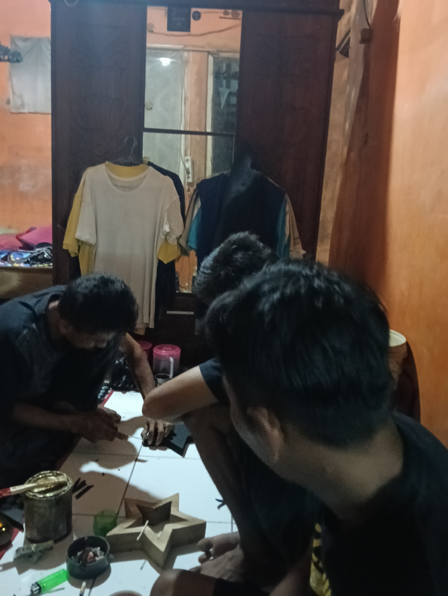 In Tasikmalaya, Indonesia, a group of individuals is engaged in a crafting activity on the floor of a dimly lit room. Three people are visible, hunched over a white tiled surface, with their backs to the camera. Their hands are actively working on a star-shaped wooden object. One person on the left is holding a small tool and appears to be applying something to the star. Another person in the center is also focused on the crafting process, while the person on the right is mostly obscured.
Scattered around the crafting area are various objects related to the activity: a can with a brush sticking out, likely containing paint or glue; a small ash tray with cigarette butts; a green lighter; and other small items. In the background, a large wooden wardrobe stands against an orange wall. Several shirts are hanging on a rack in front of the wardrobe, including a white t-shirt and a yellow t-shirt. The overall environment suggests a casual, perhaps informal workshop setting.