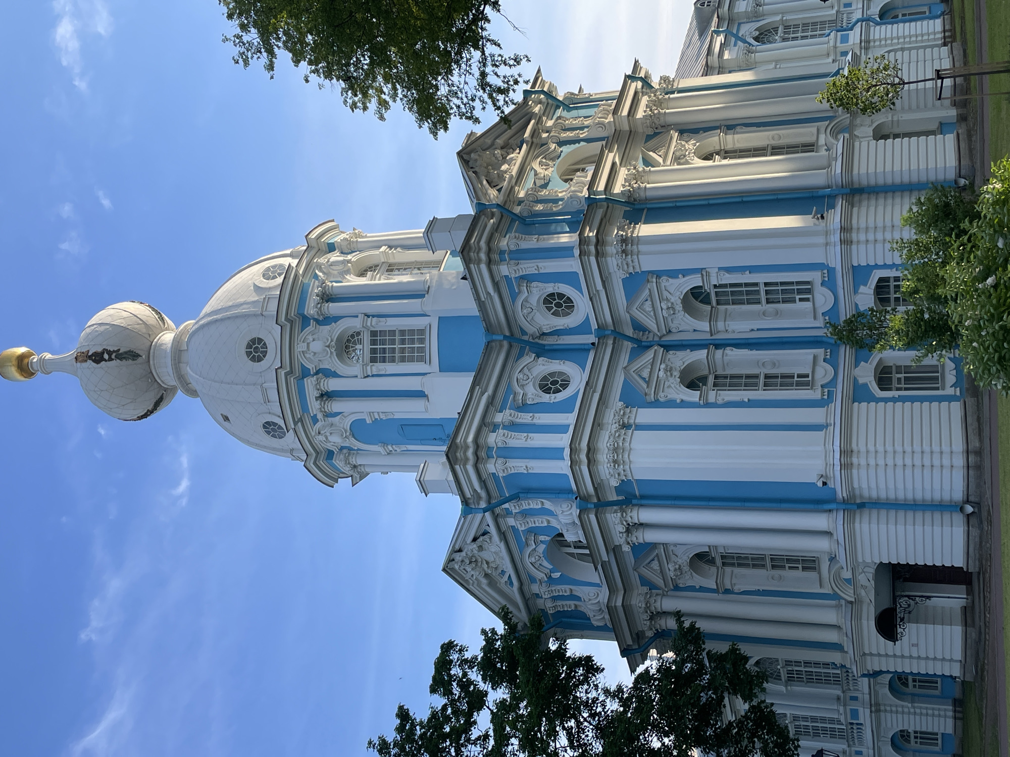 This image captures the Smolny Cathedral (Smolny Convent of the Resurrection) located in St. Petersburg, Russia. The large, ornate building occupies the majority of the frame, presented from a slightly low-angle perspective, emphasizing its height and grandeur. The church exhibits a distinct Baroque architectural style, characterized by its vibrant light blue and white color scheme.
The facade is intricately detailed with numerous white Corinthian columns and pilasters, often grouped, framing tall arched windows with multi-pane glazing. Decorative elements such as sculpted pediments, friezes, and scrolls adorn the upper sections of the walls. A prominent white central dome, featuring a ribbed or paneled surface, culminates in a smaller white cupola. This cupola is adorned with dark, stylized figures or bas-reliefs around its base and is topped by a golden spherical finial. Blue vertical downspouts are integrated into the blue sections of the building. On the lower left, a dark arched entrance with a black awning is visible. A small black security camera is mounted on a blue wall section on the right side.
The building is set against a clear, bright blue sky with a few faint wispy clouds. Strong sunlight illuminates the scene, casting highlights on the white architectural details and creating some shadowed areas on the left side of the building. Lush green deciduous trees partially frame the church on both the left and right sides. In the foreground, a landscaped area features green grass, various small green bushes, and young trees. A paved path runs along the base of the building, bordering the grassy area.